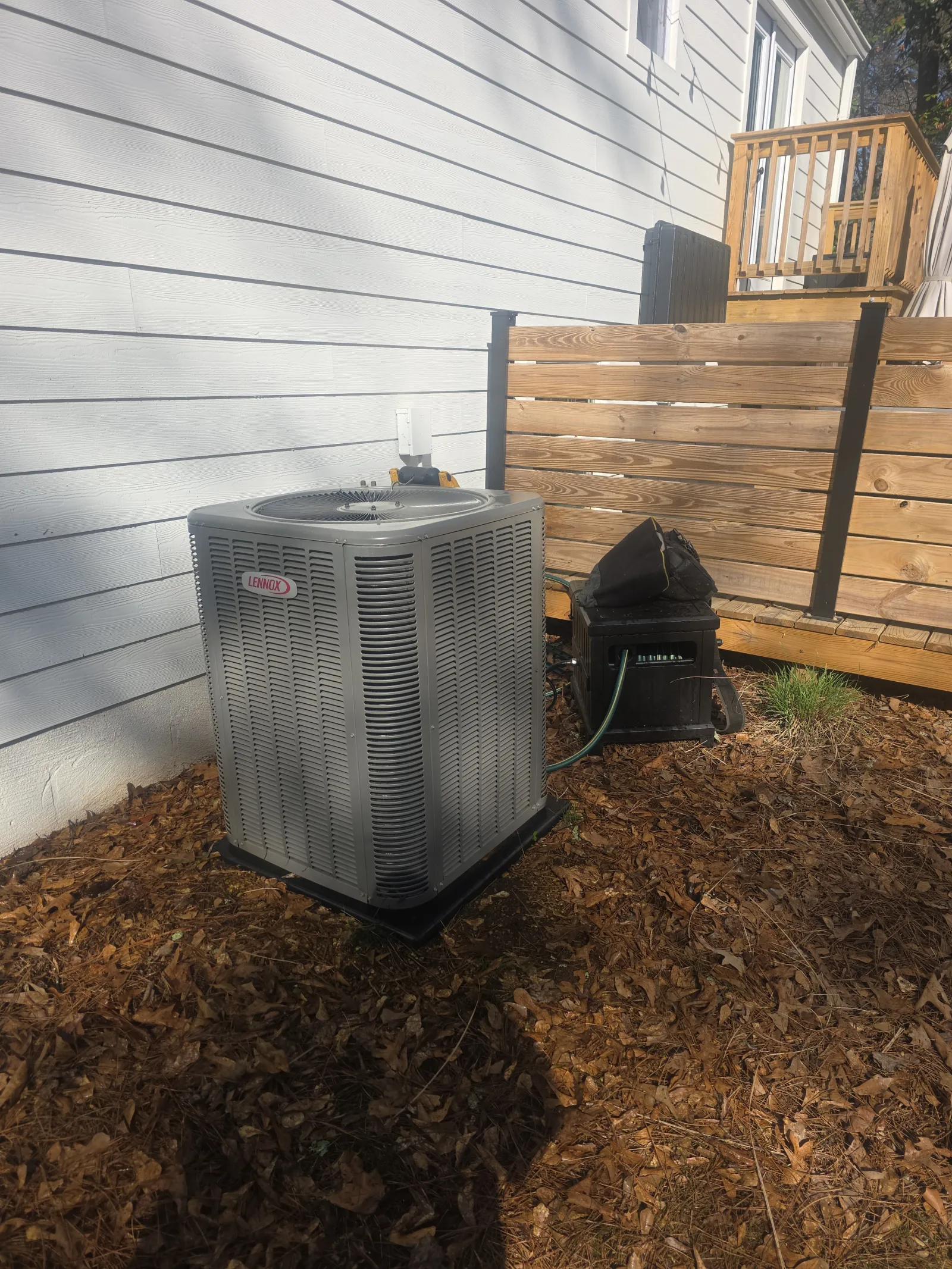 HVAC Repair, Maintenance and Installation in Roswell by The Air Company of GA. Outdoor Lennox air conditioning unit next to white house wall and wooden fence in a yard covered with dry leaves.