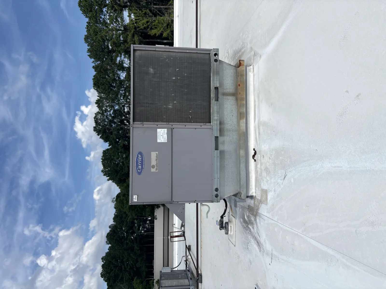 Commercial rooftop HVAC unit on white roof under blue sky with scattered clouds and trees in background.