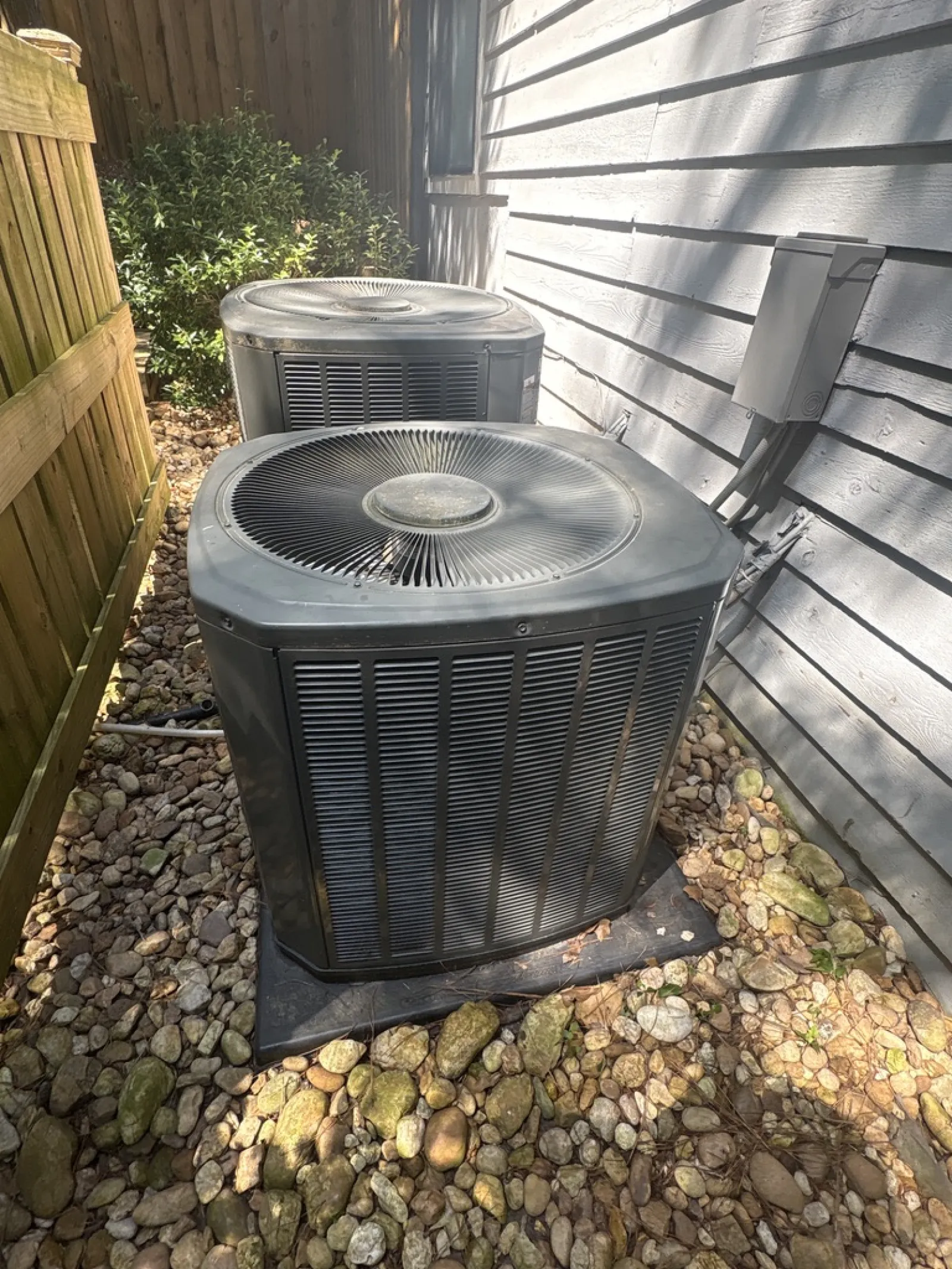 Two outdoor air conditioning units on a bed of rocks next to a house and wooden fence in sunlight.