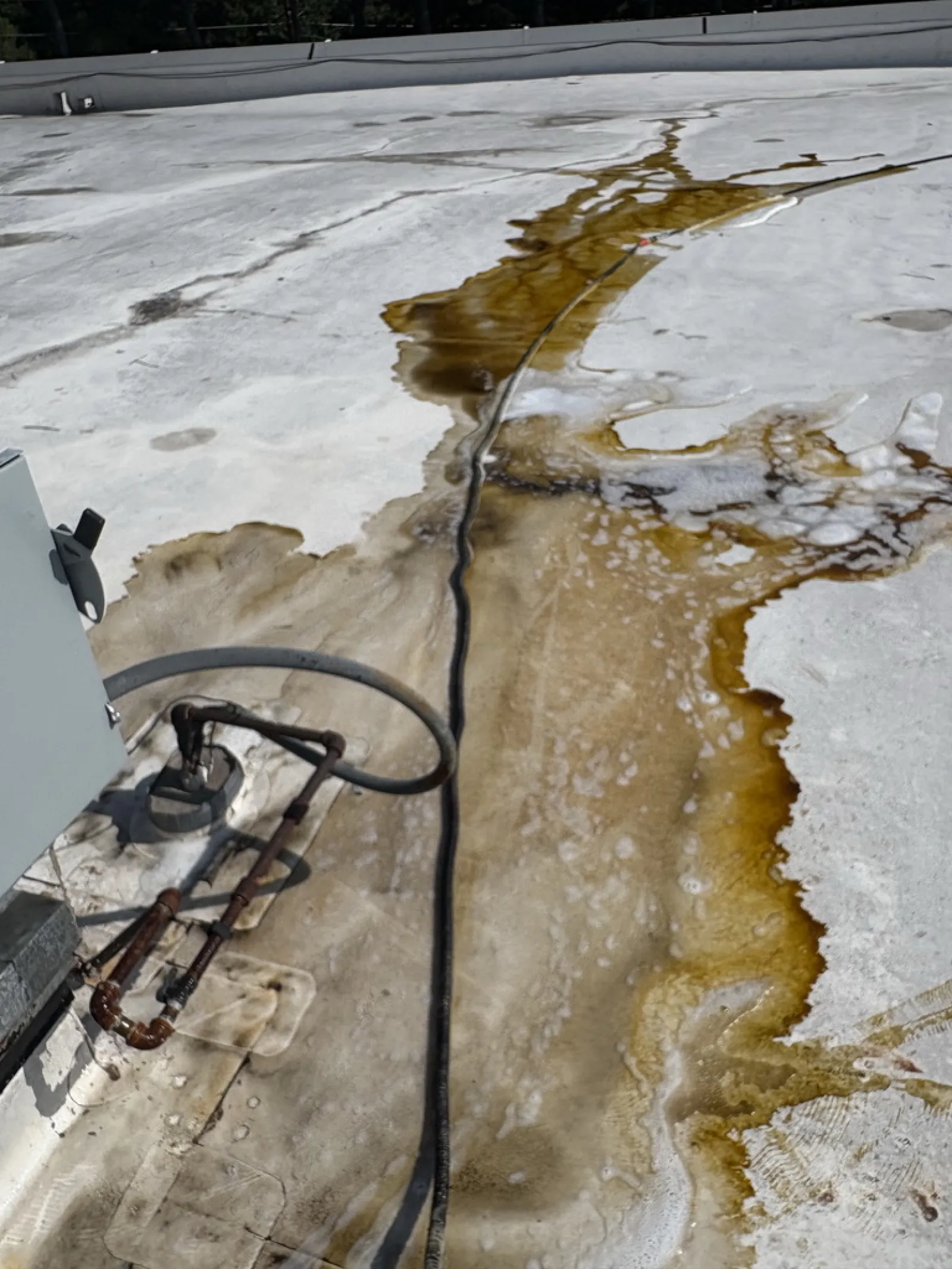Rusty pipes and a black cable on a dirty white rooftop with large brown water stains and patches.