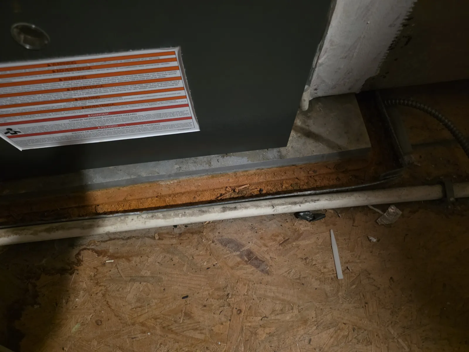 Close-up of a furnace base with warning label and a white pipe on a wooden floor in a dimly lit utility area.