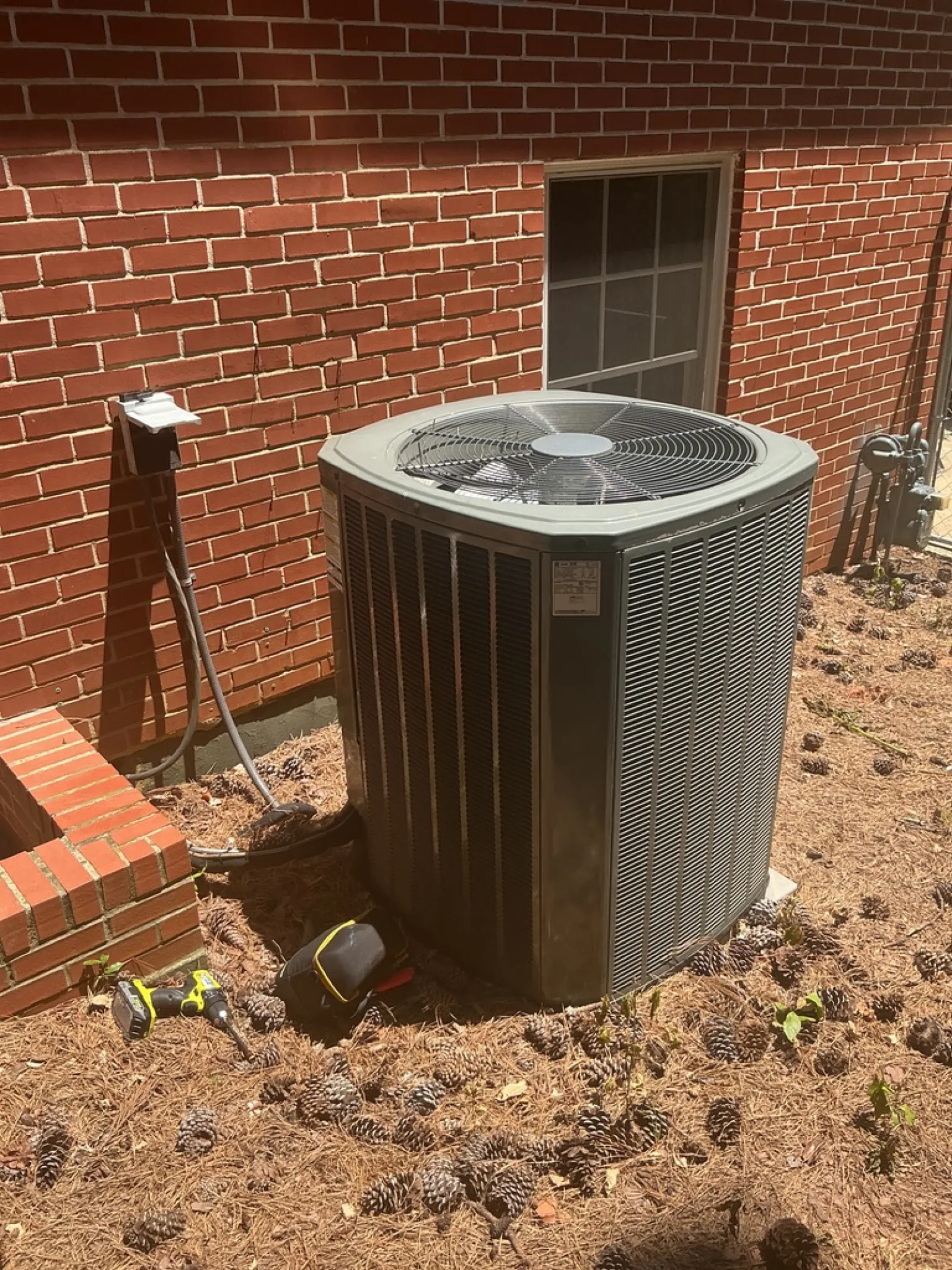 Outdoor air conditioning unit installed next to brick house with tools and pinecones nearby on ground.