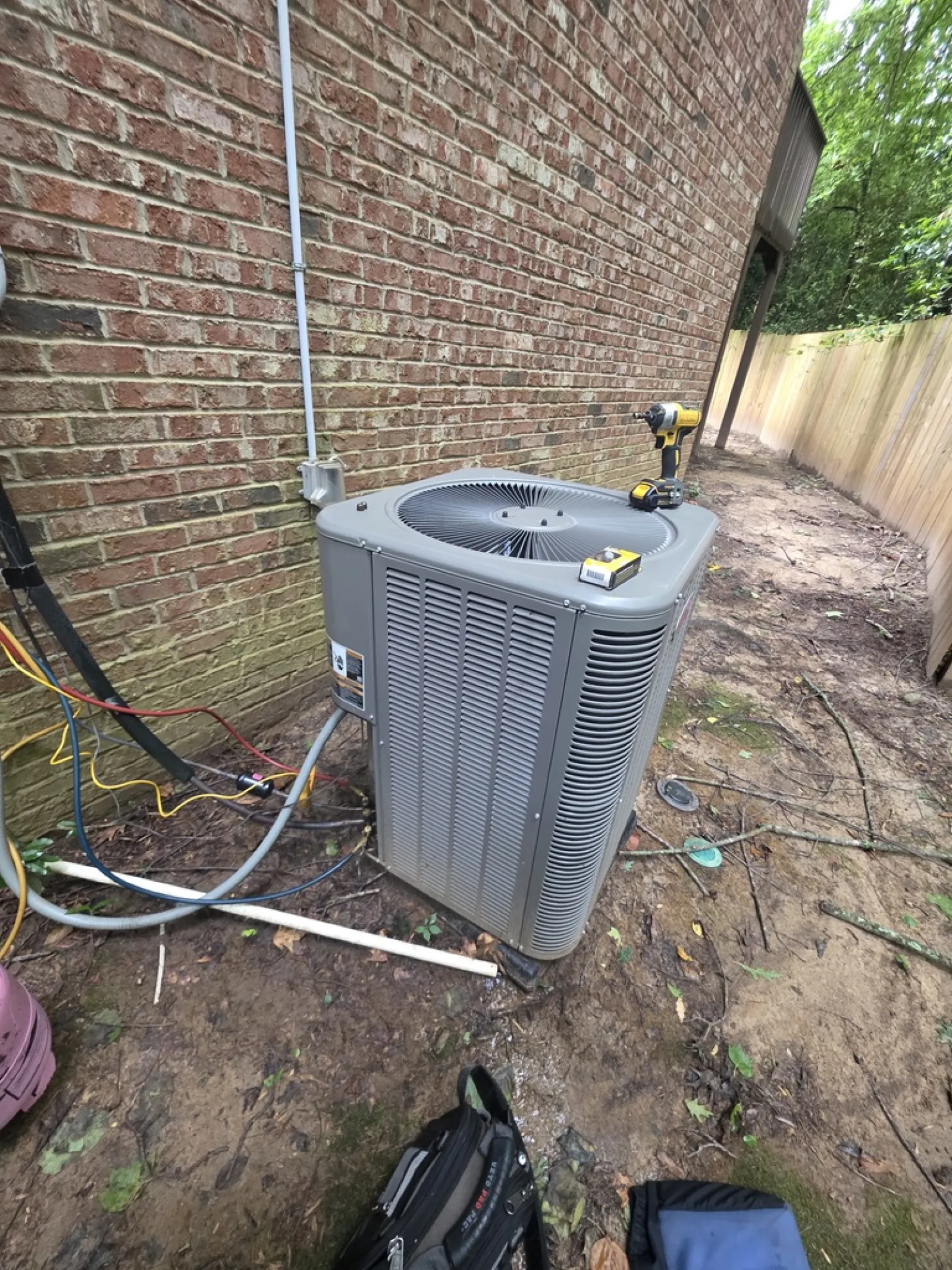 Outdoor air conditioning unit installed next to a brick wall with tools and wiring around it in a backyard.