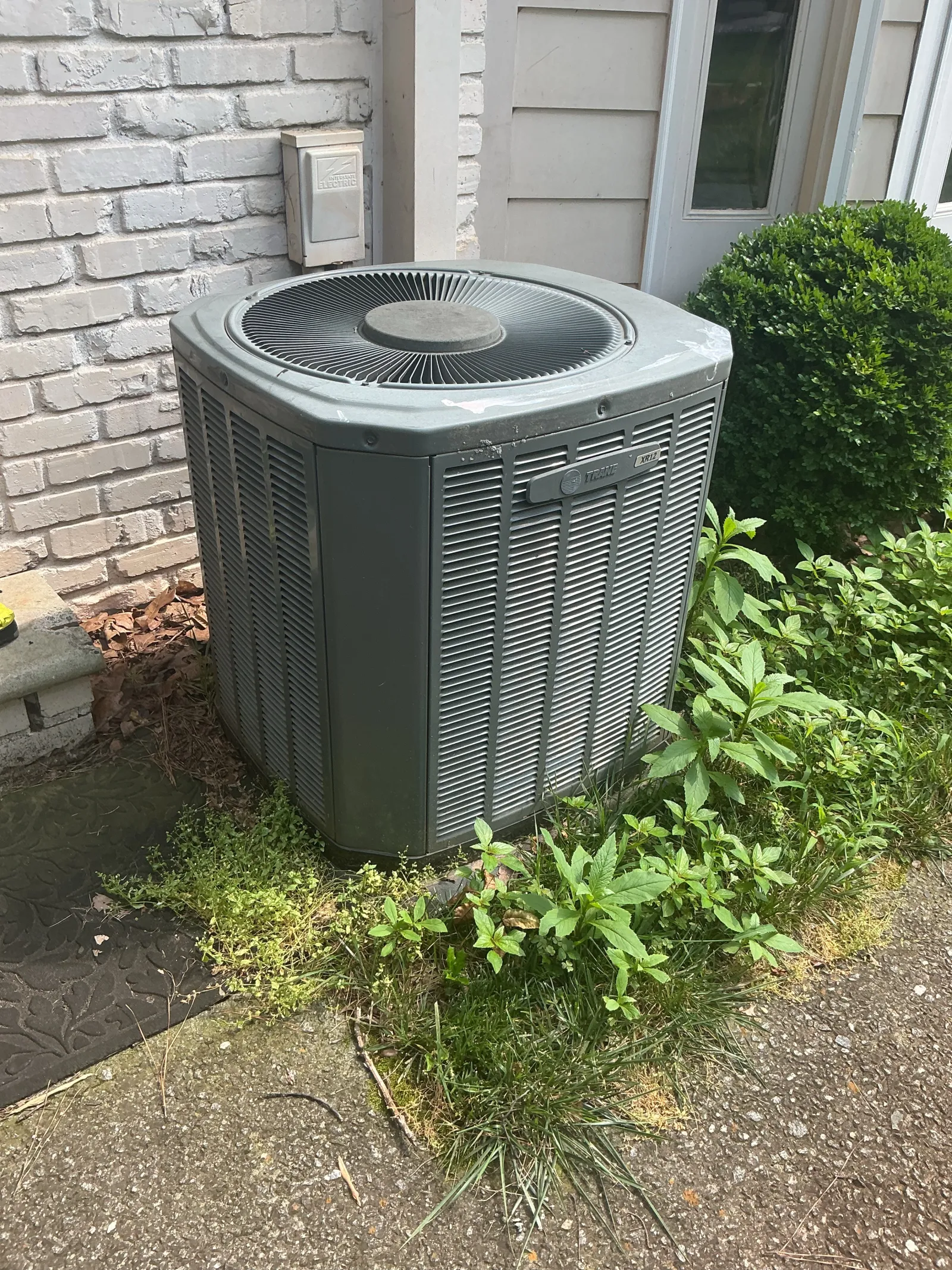 Outdoor residential air conditioning unit beside a white brick wall with greenery and shrubs nearby.