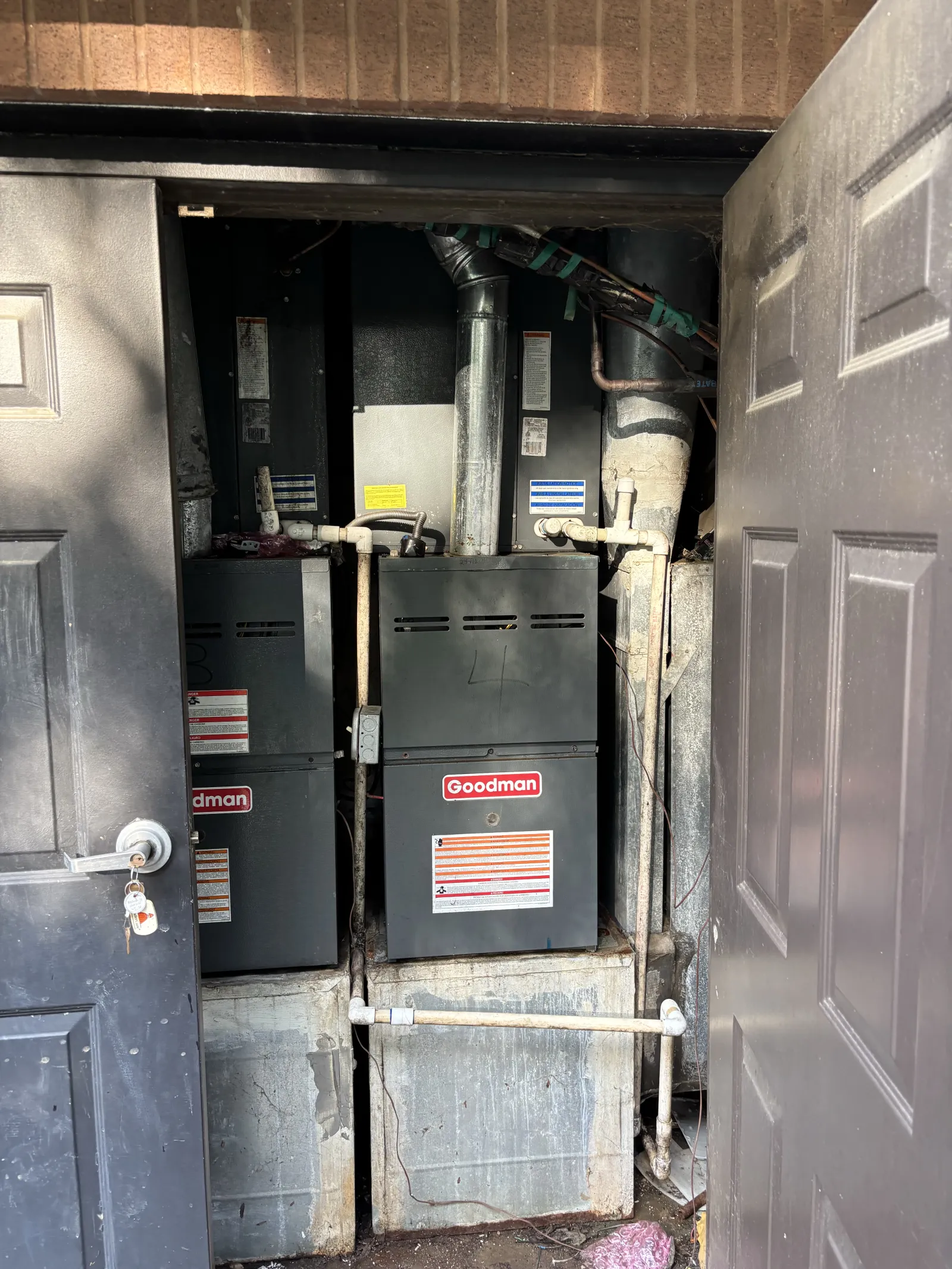 Two Goodman HVAC units installed in a utility closet with open double doors and exposed pipes.