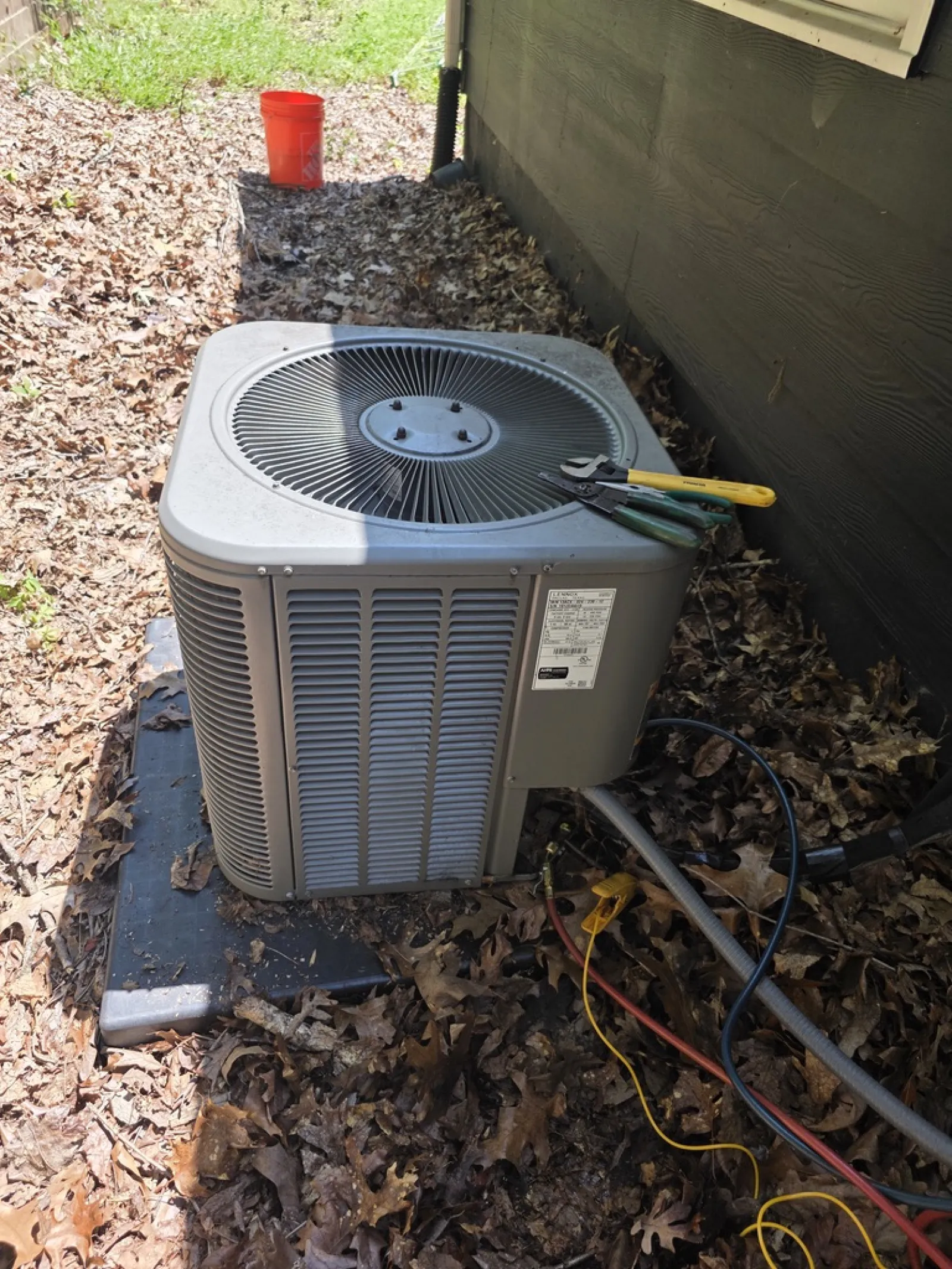 Outdoor air conditioning unit on a concrete pad with tools and cables, surrounded by dry leaves near a house wall.