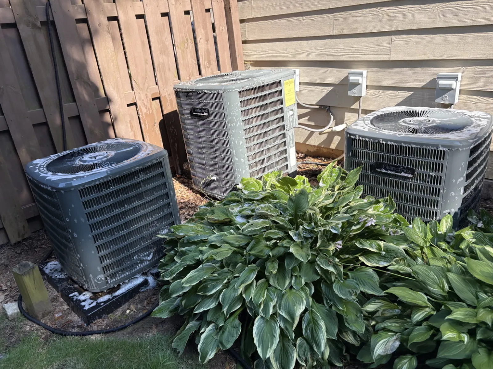 Three outdoor air conditioning units behind a house next to green leafy plants and wooden fence on a sunny day