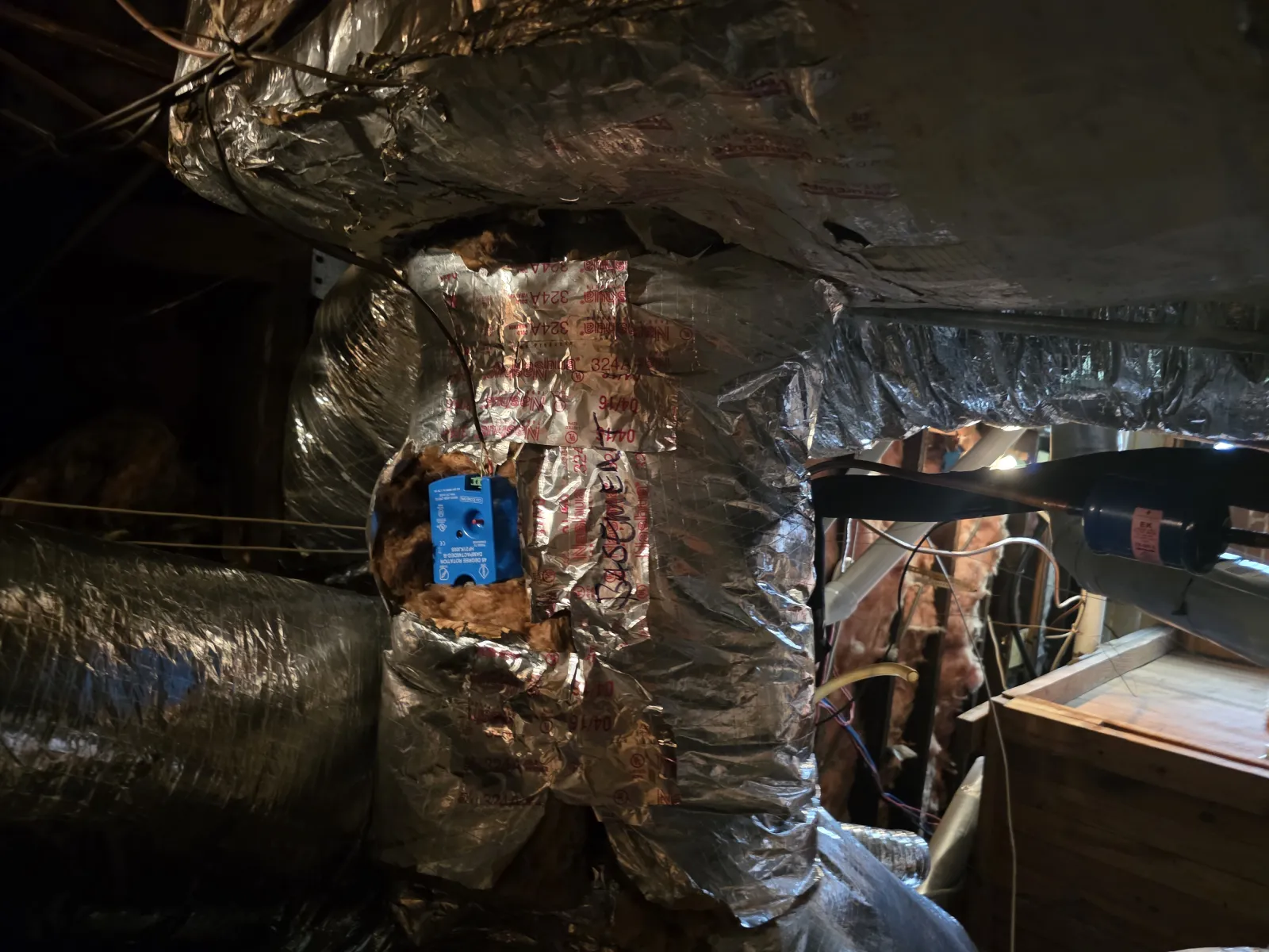 Foil-insulated HVAC duct with exposed blue control box and surrounding wires in a dimly lit basement.