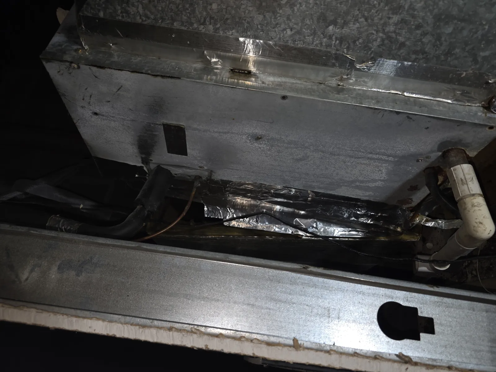 Close-up of an HVAC unit's metal components showing ductwork, insulation, and a white PVC drain pipe.