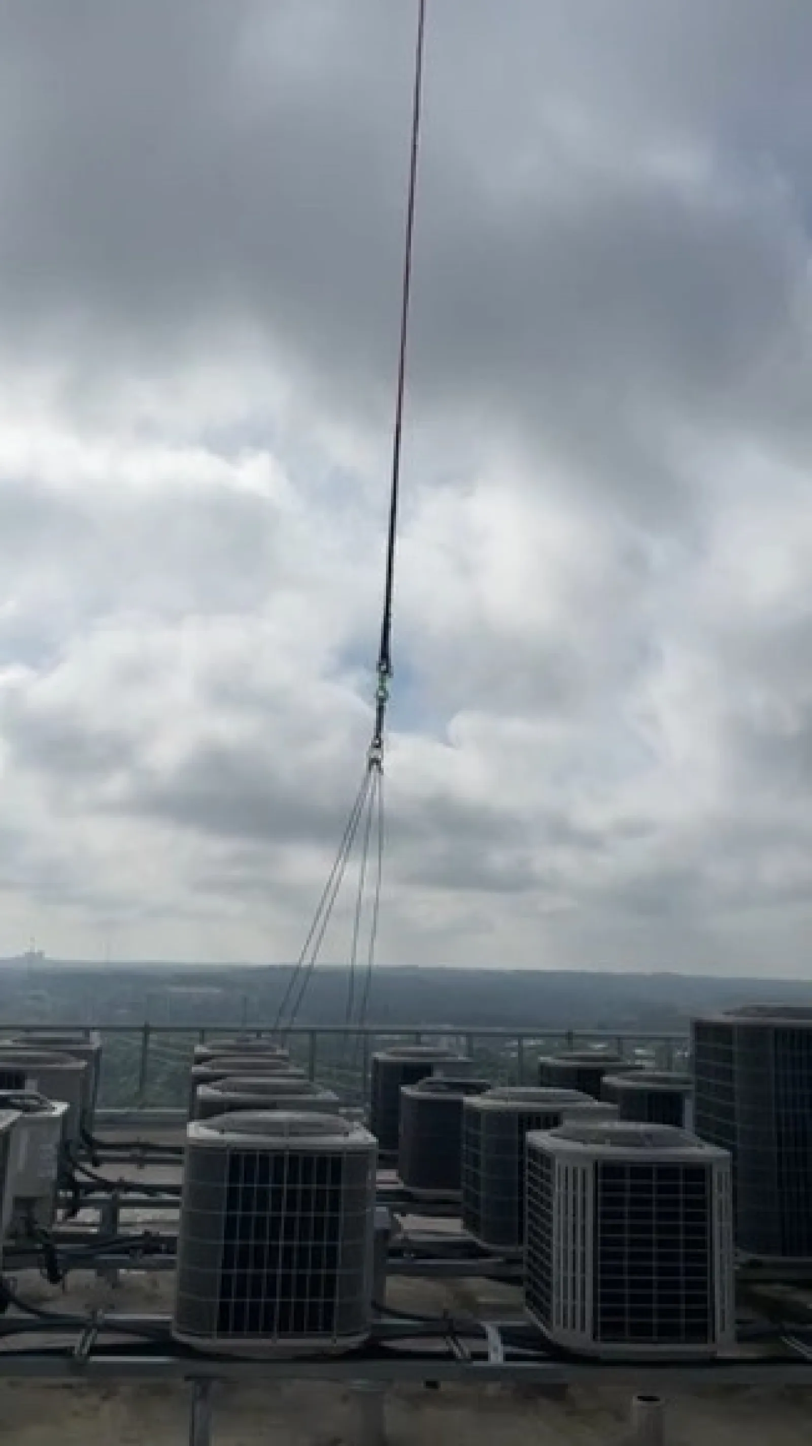 Crane lifting HVAC unit above rooftop with several air conditioning units under cloudy sky