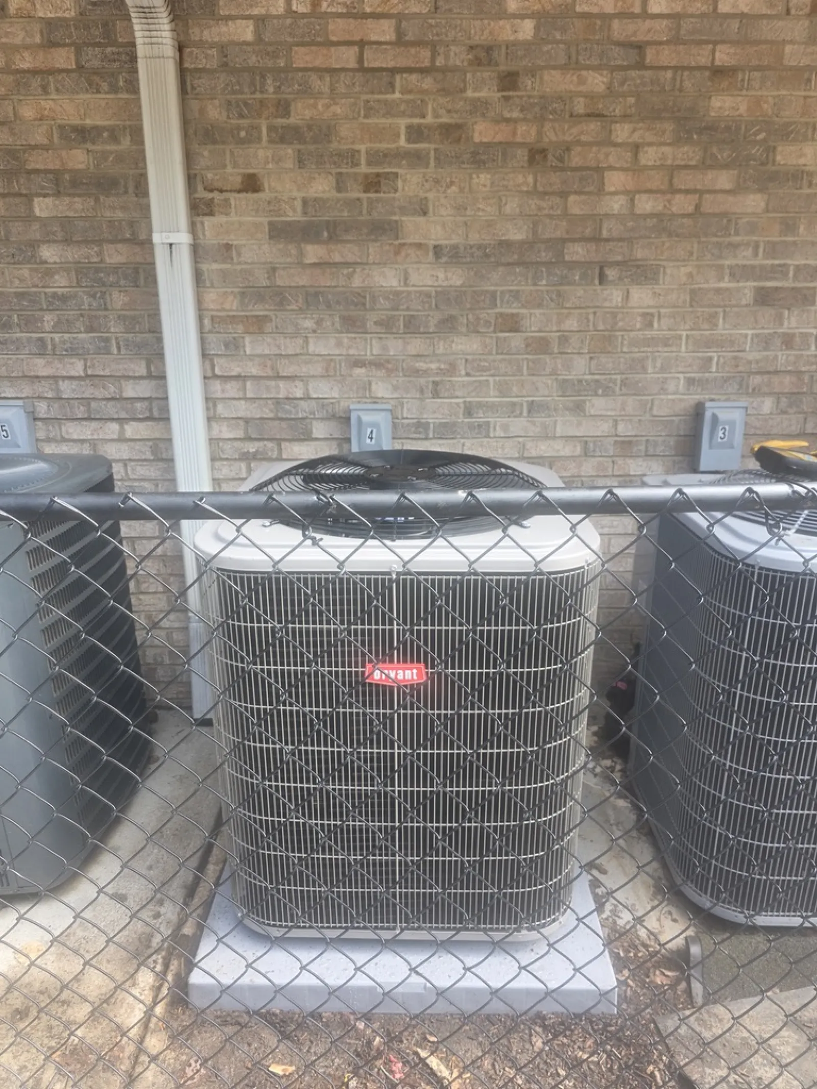 Three HVAC units behind a chain-link fence against a brick wall with numbered electrical boxes above.