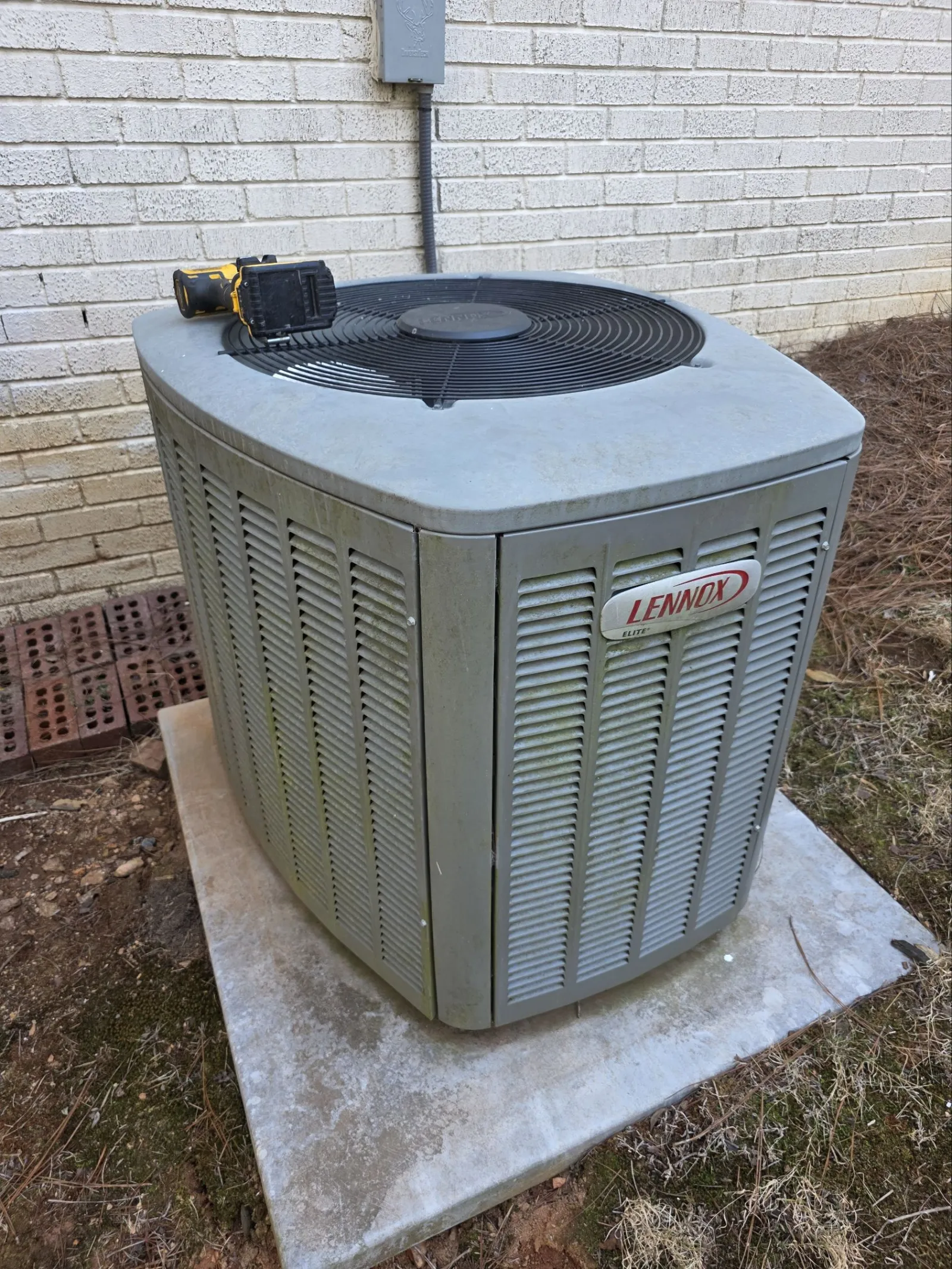 Residential AC Cooling Repairs in Sandy Springs 30350 by The Air Company of GA. Outdoor Lennox Elite air conditioning unit on concrete pad next to brick house with cordless drill on top