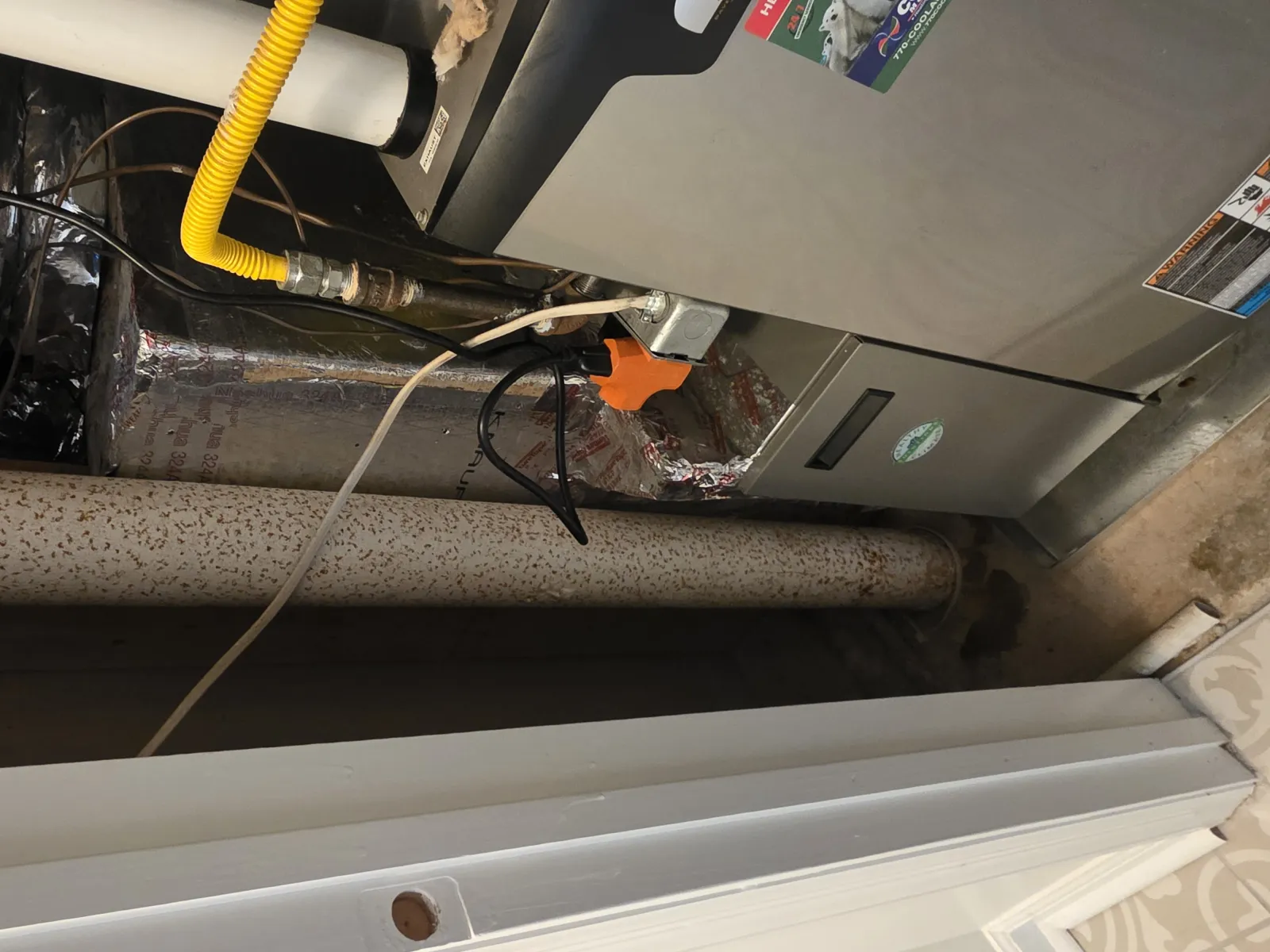 Basement furnace with yellow gas line, insulated ducts, and rusted pipe in a utility space near tiled floor.