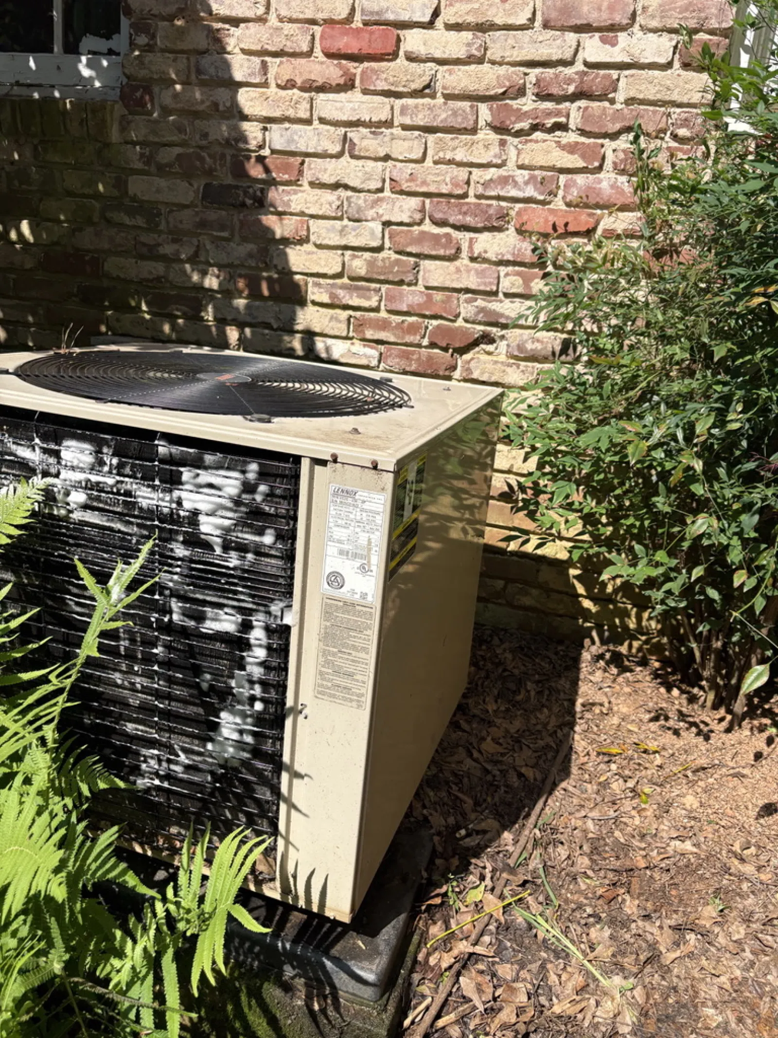 Outdoor air conditioning unit beside brick wall with surrounding plants and greenery in sunlight.
