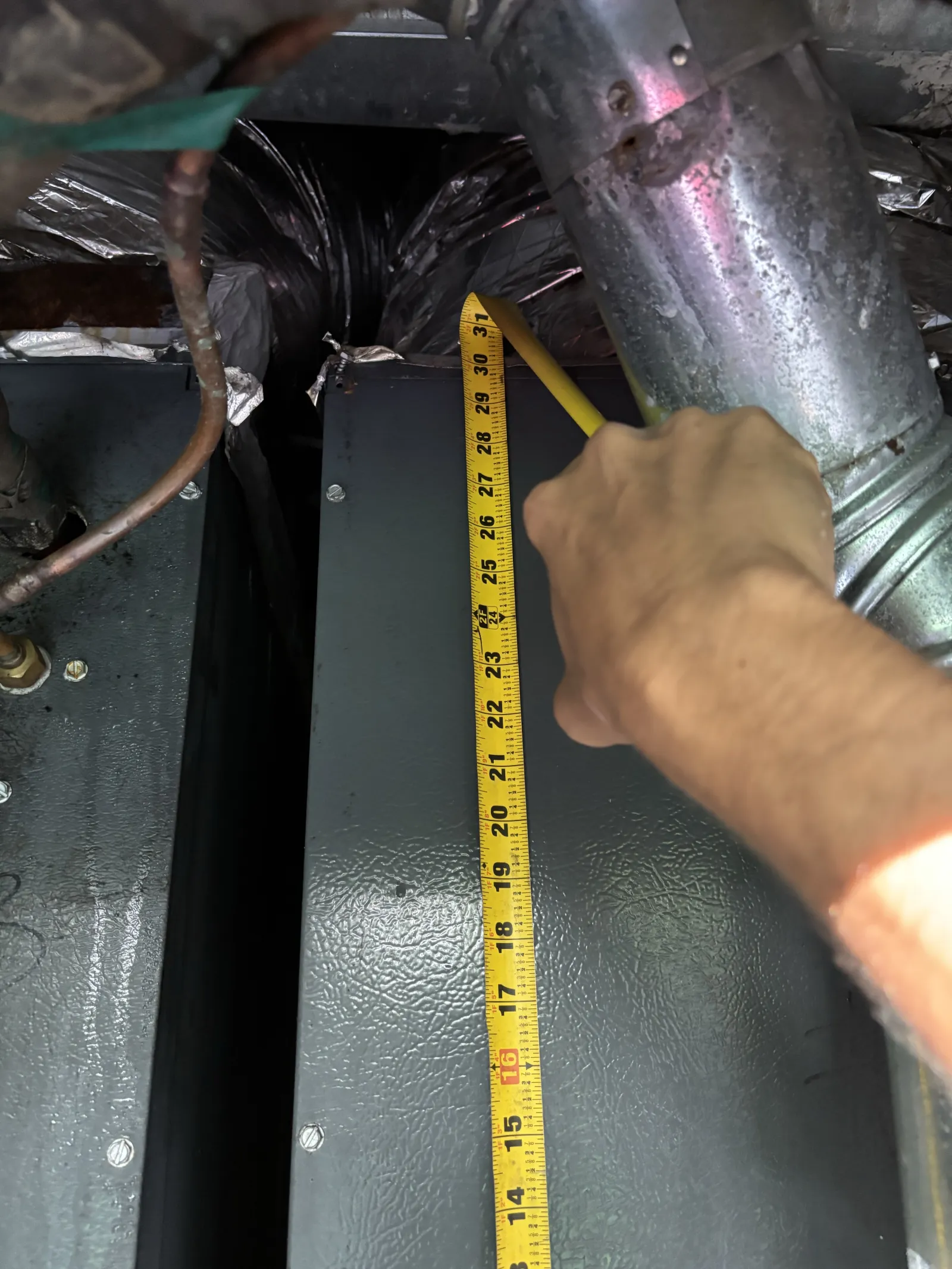 Hand measuring length of metal panel with a yellow tape measure under ductwork in an industrial setting.