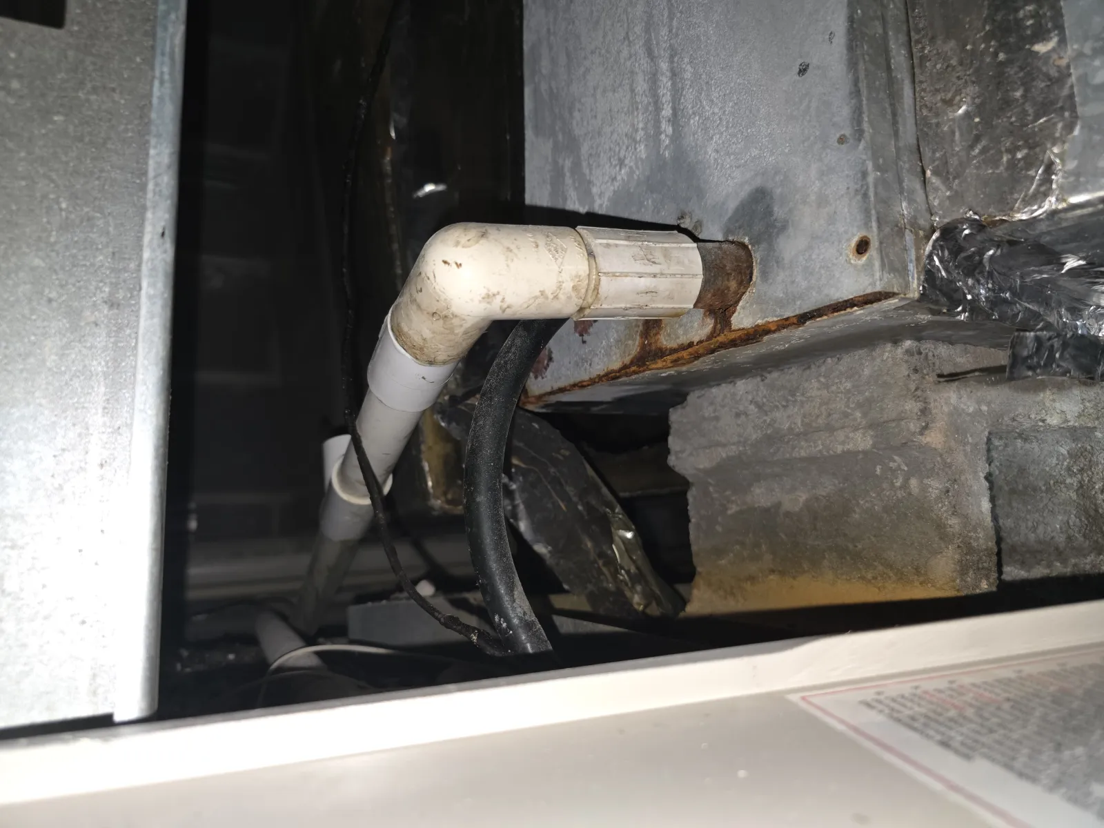 Close-up of rusted HVAC duct with connected white PVC pipe and black cables in a basement or utility area