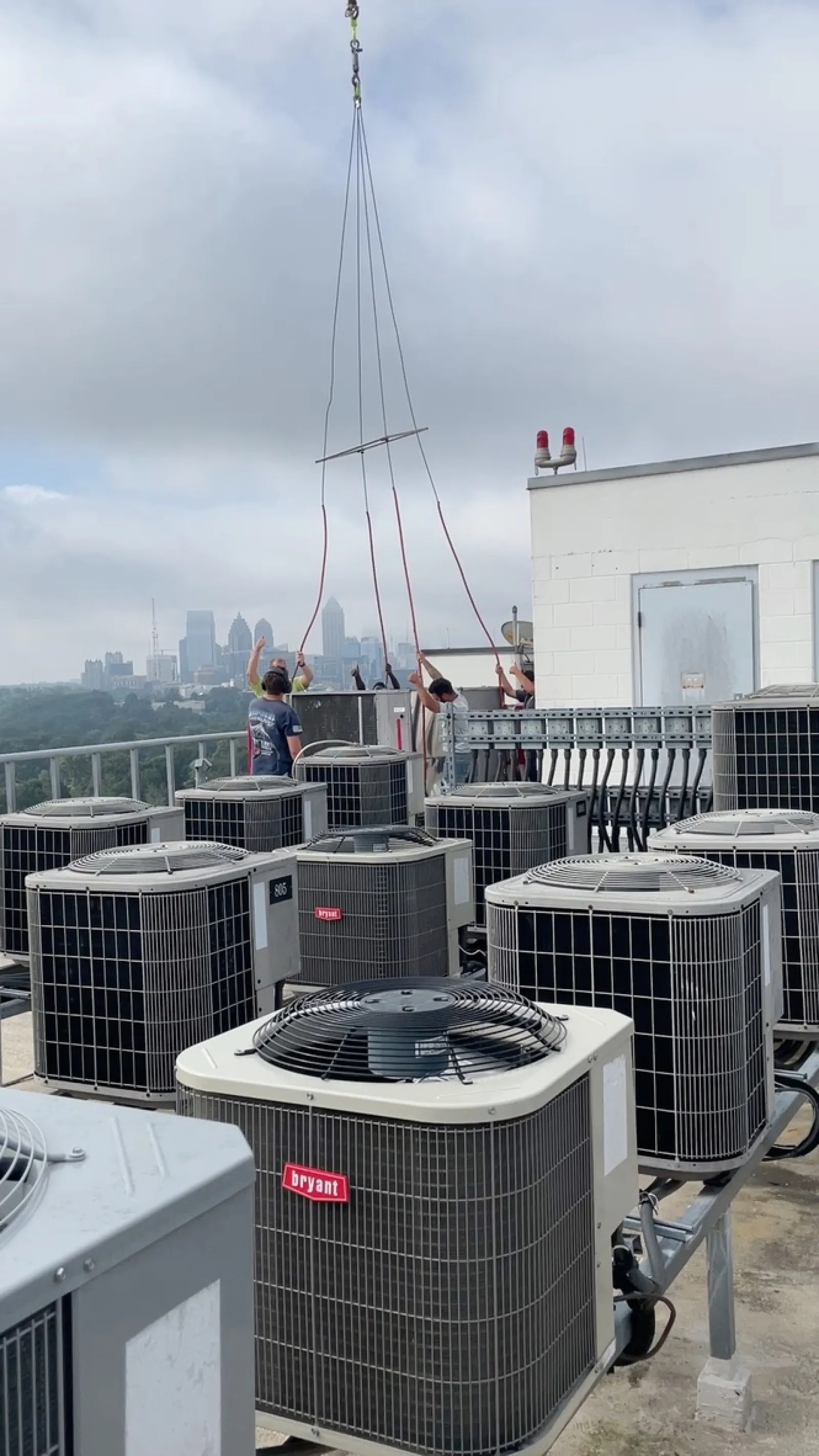 Technicians installing HVAC units on a rooftop with city skyline under a cloudy sky.