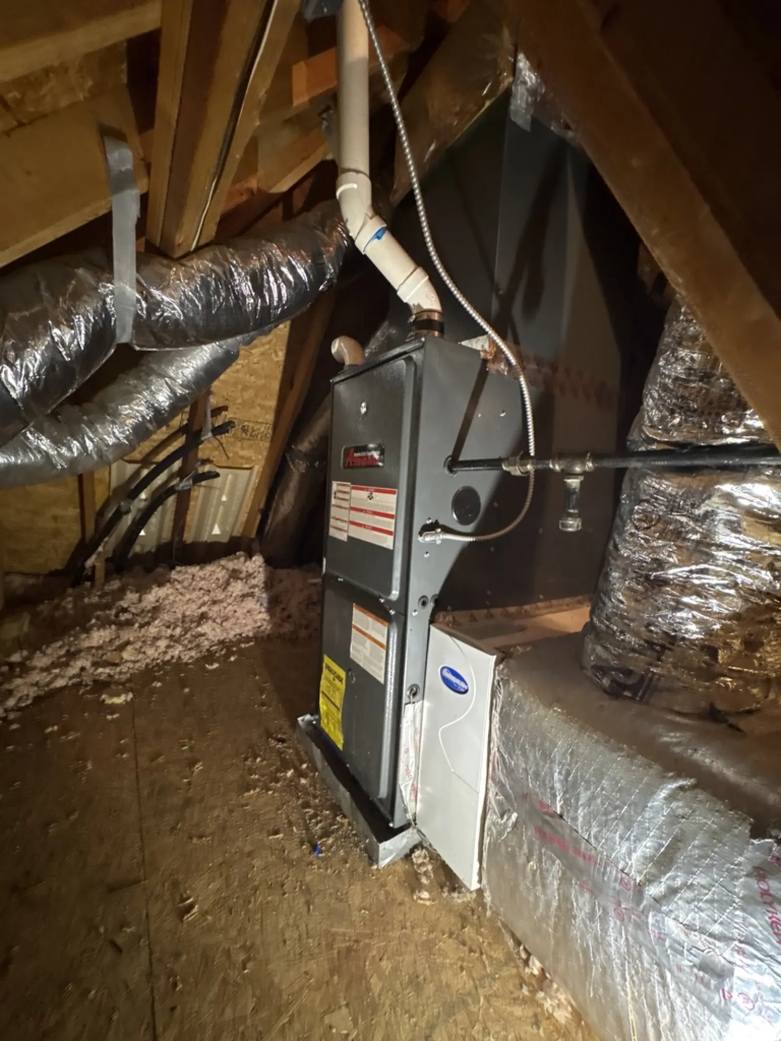Furnace and HVAC ductwork installed in a wooden attic with insulation and exposed beams.