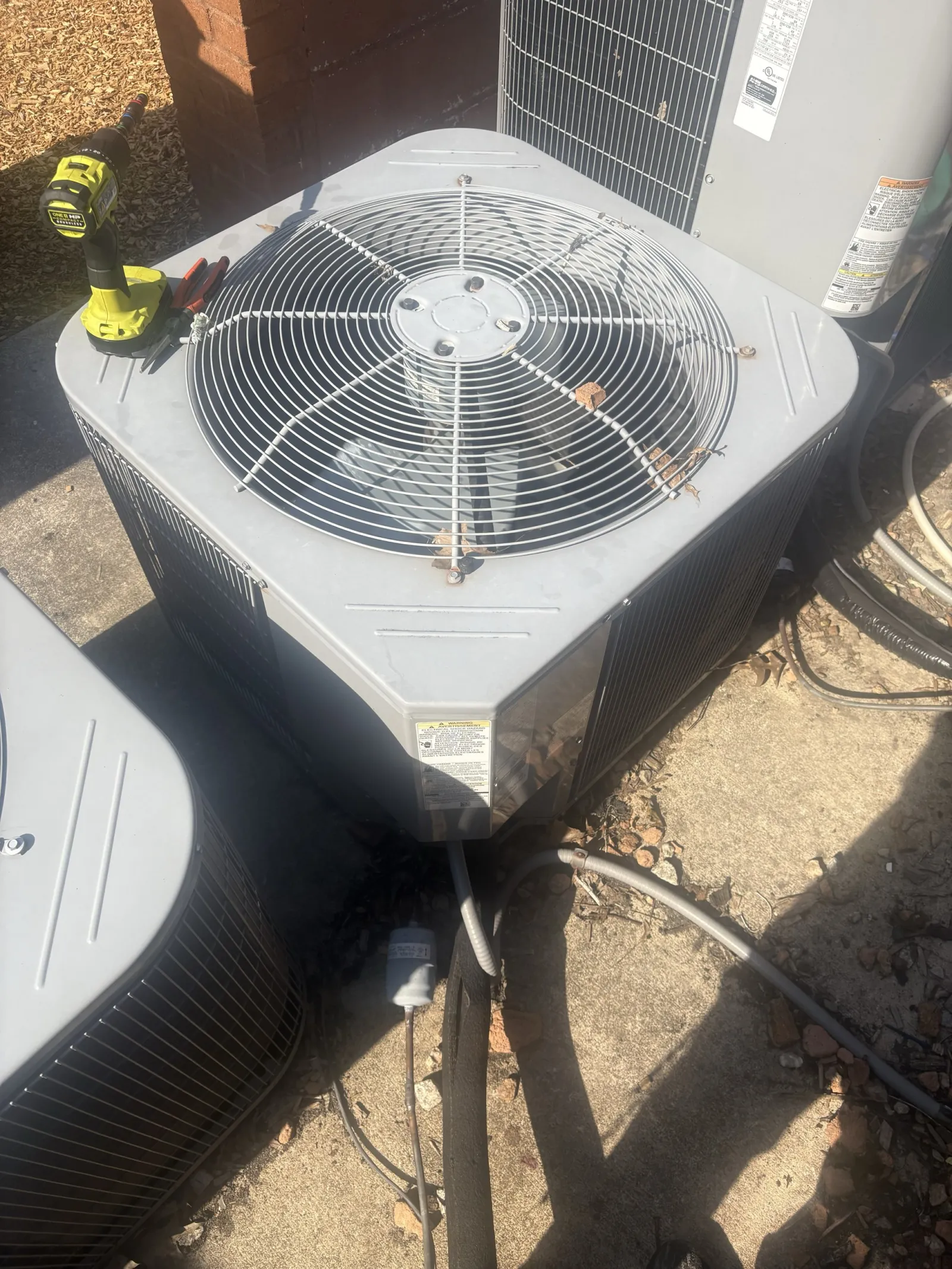 Outdoor HVAC unit with protective grill and power drill on top, placed beside another air conditioning unit.