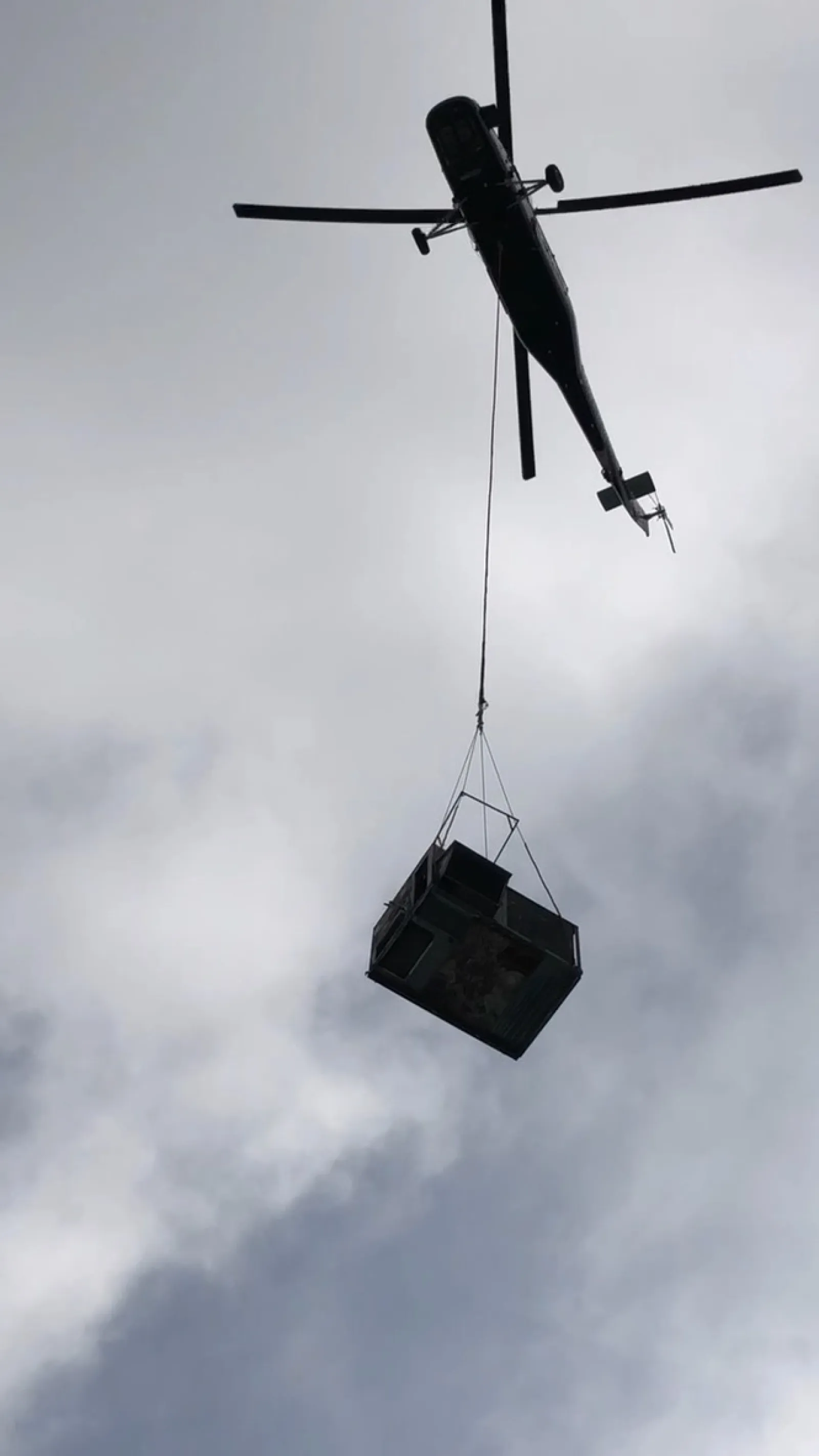 Helicopter lifting a large metal container high against a cloudy sky during aerial transport.