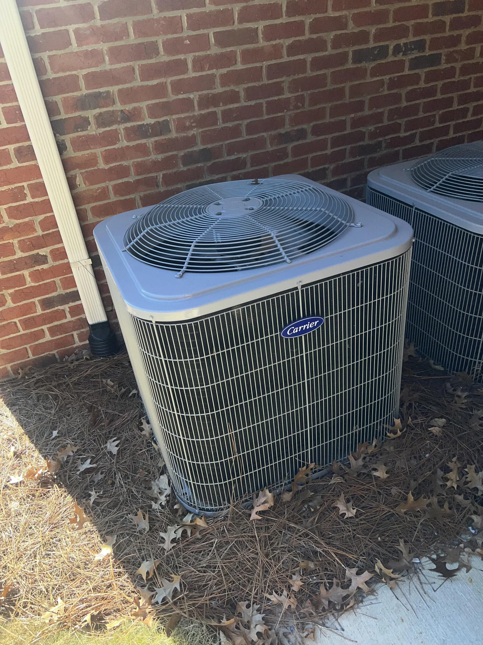 Atlanta AC Cooling Air Conditioner Repair in Vinnings 30339 by The Air Company of GA. Two Carrier outdoor air conditioning units next to a brick wall with dried leaves and pine straw on the ground.