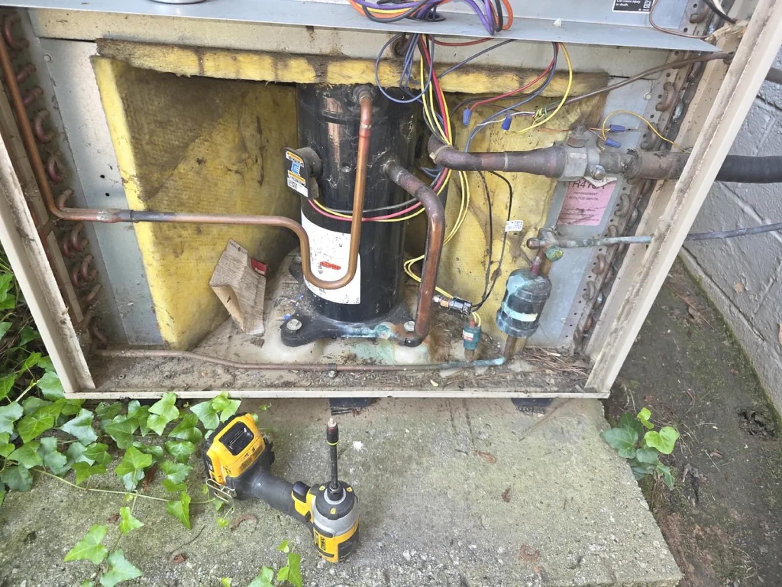 Open HVAC unit showing compressor, copper pipes, wiring, insulation, and a yellow power drill on the ground.
