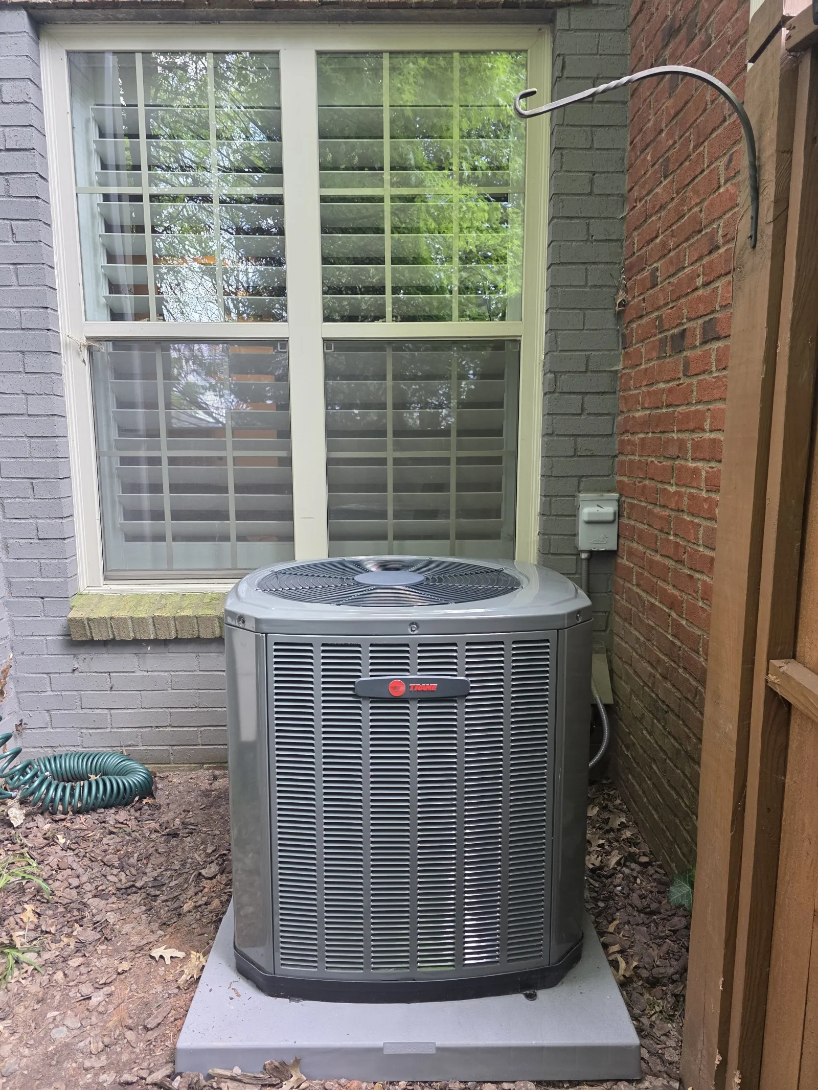 Outdoor air conditioning unit installed below a double window between brick and wooden walls with garden hose nearby