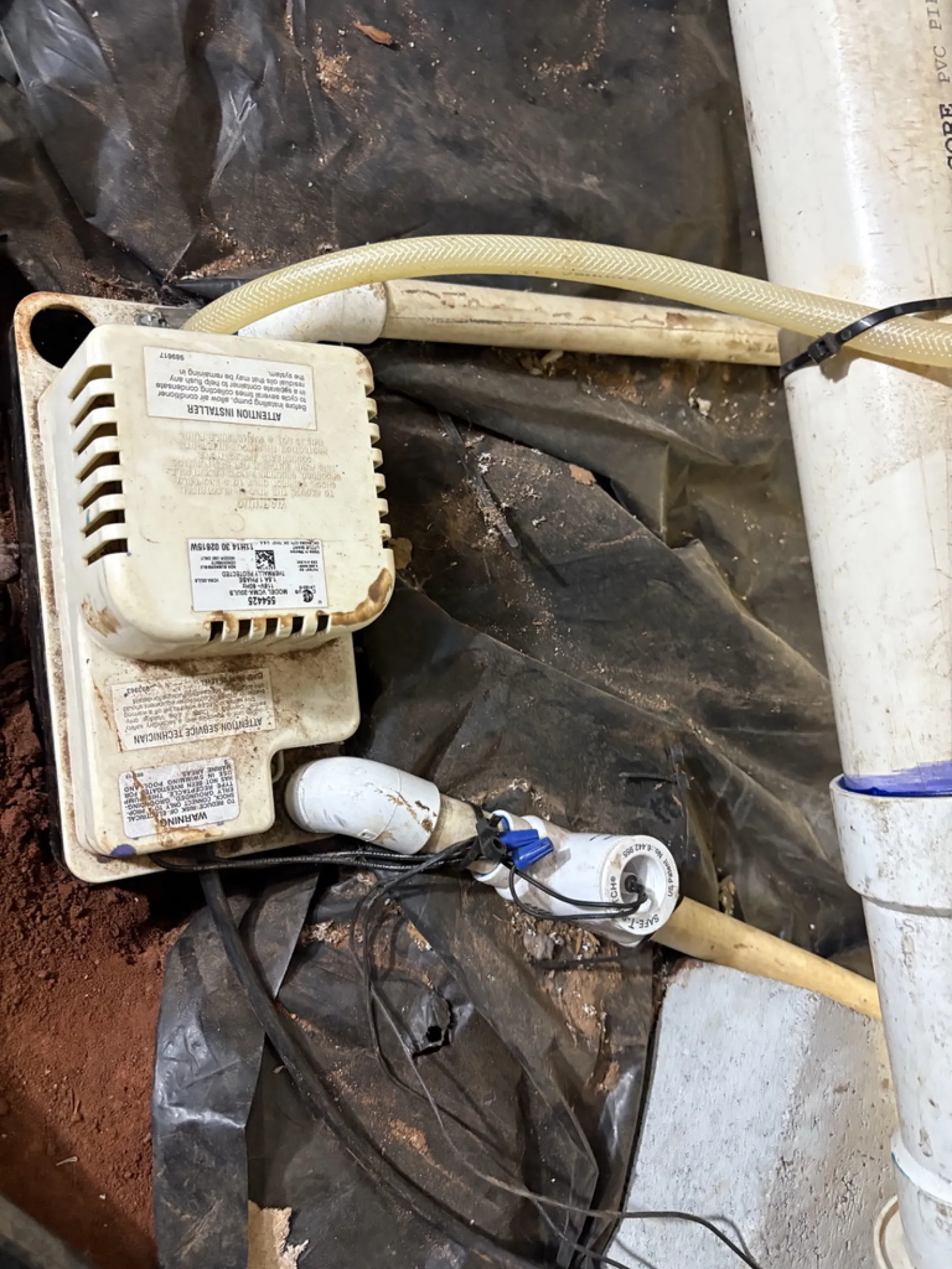 Old water pump connected to white PVC pipes and black plastic liner on dirt floor in a utility area.