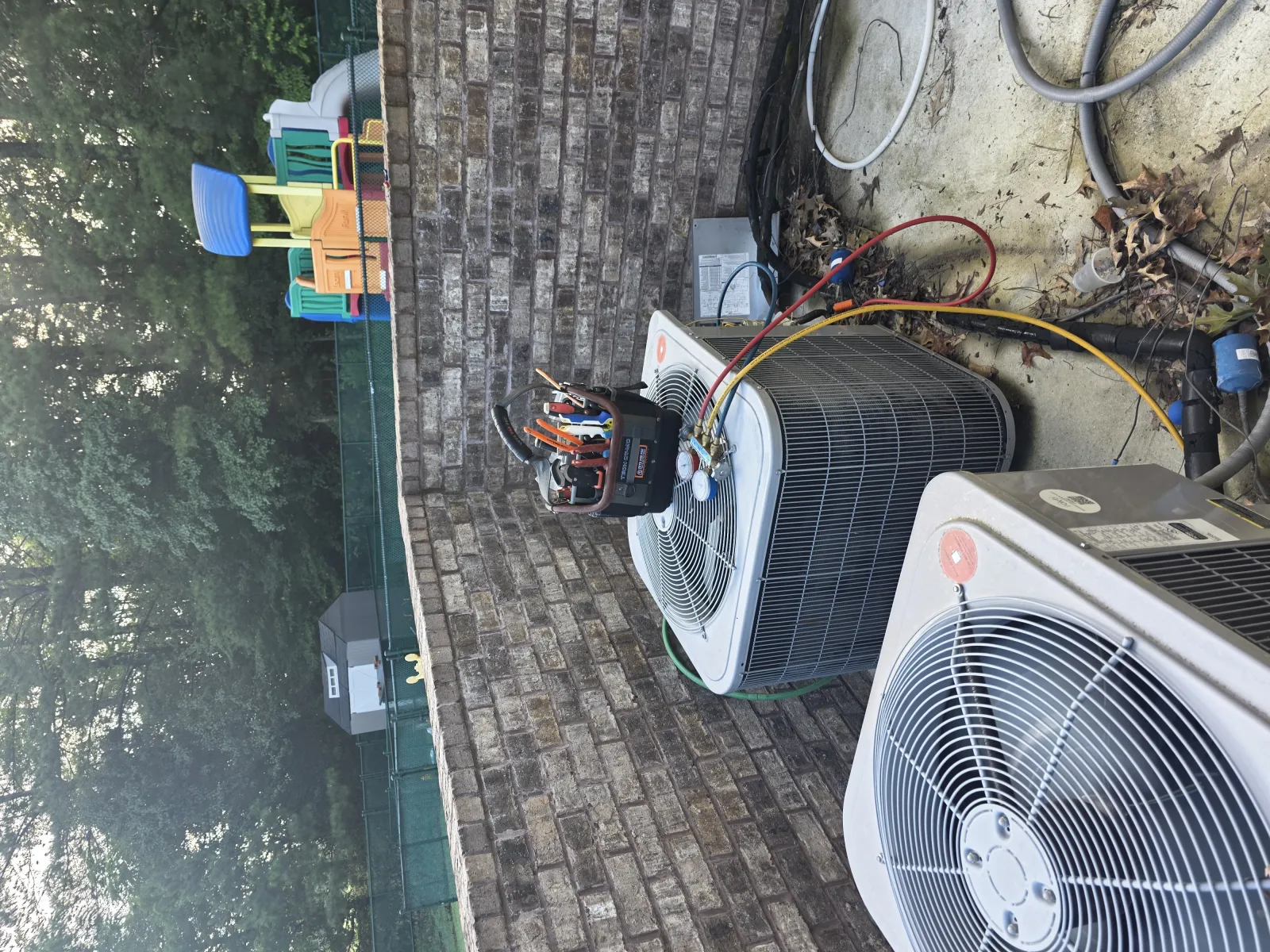 Two outdoor air conditioning units with diagnostic tools attached against a brick wall in a backyard.