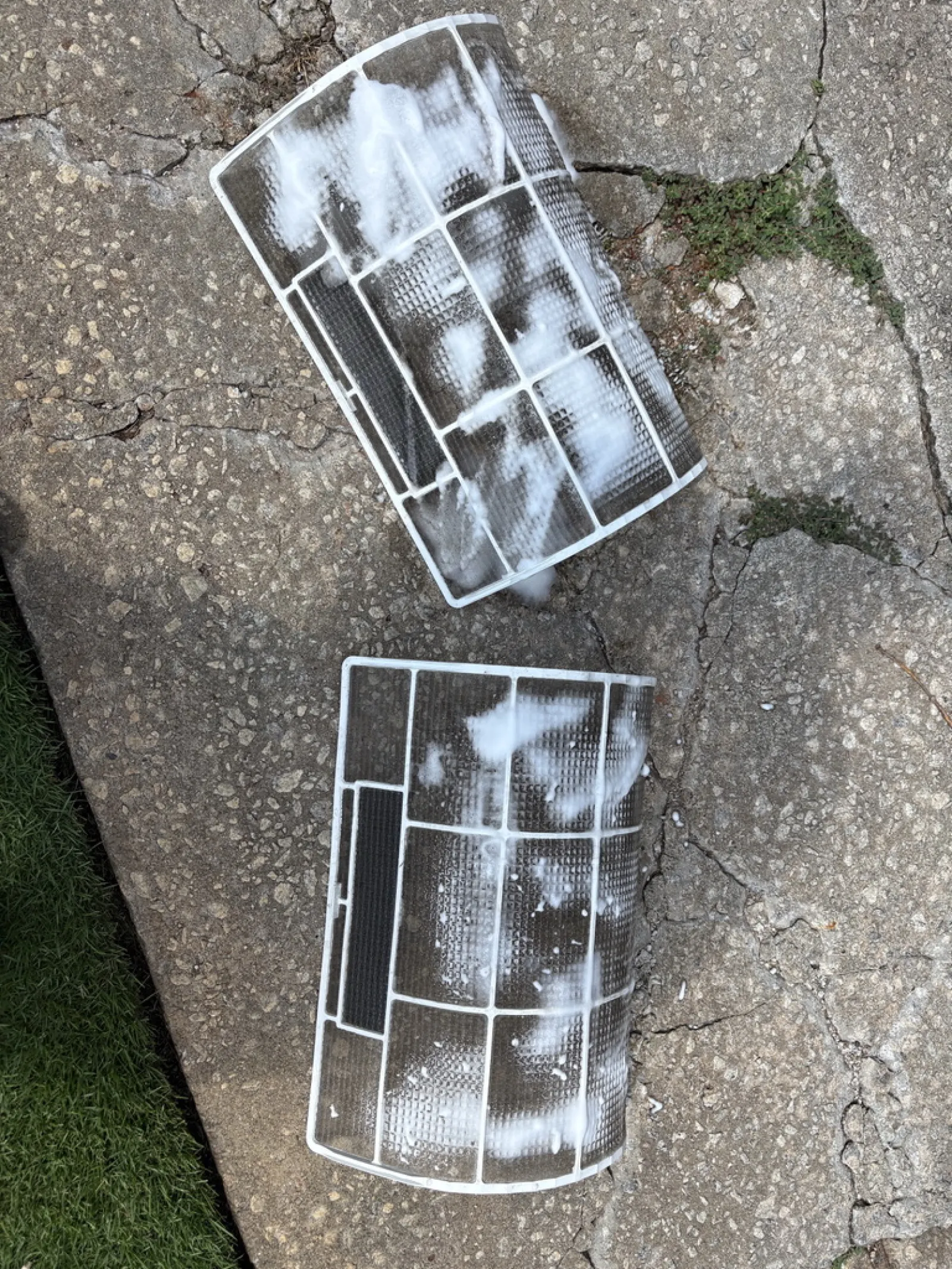 Two rectangular air conditioner filters with soap suds resting on cracked concrete pavement next to grass.