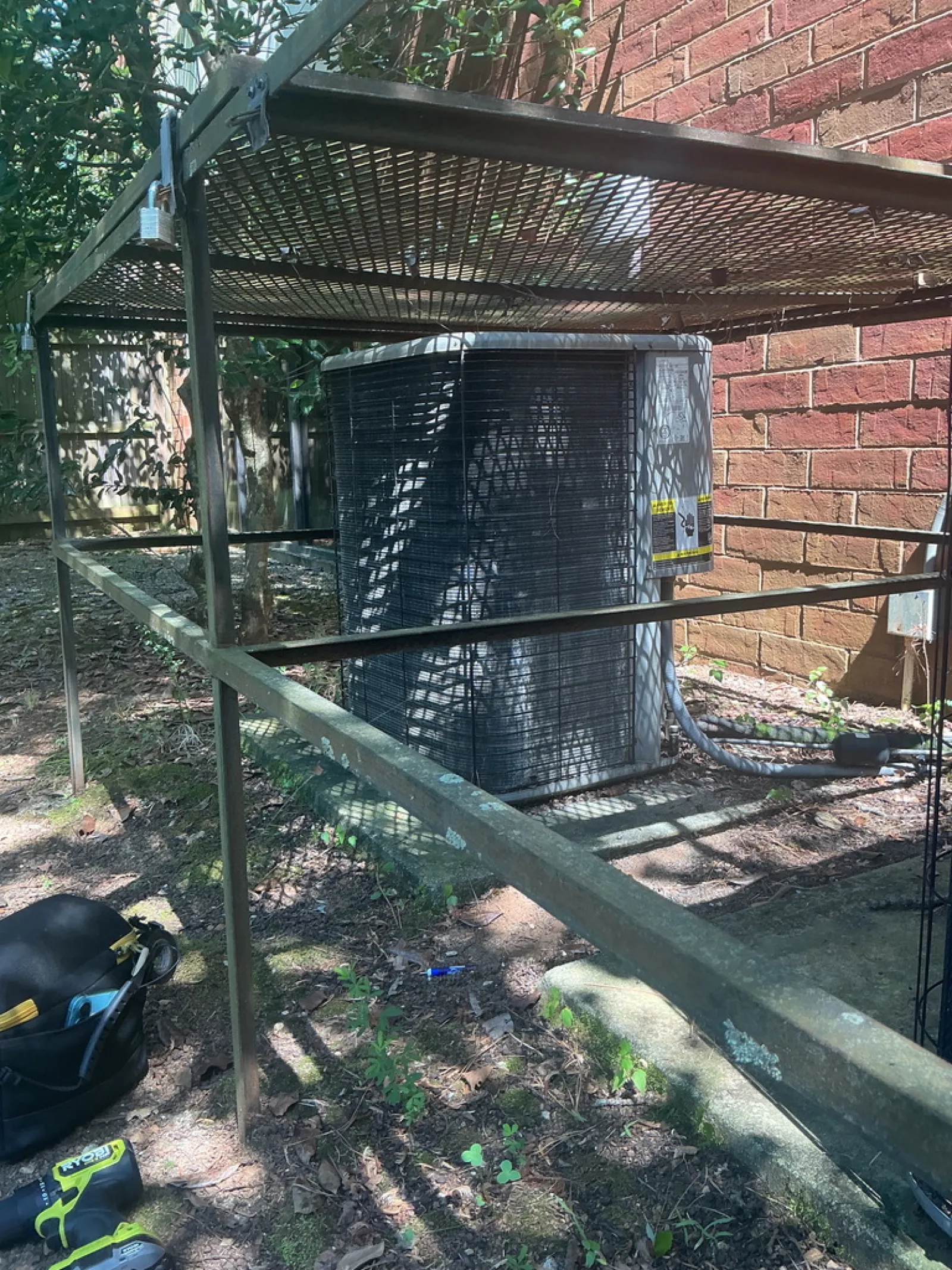 Outdoor air conditioning unit protected by a metal cage beside a brick wall with tools on the ground