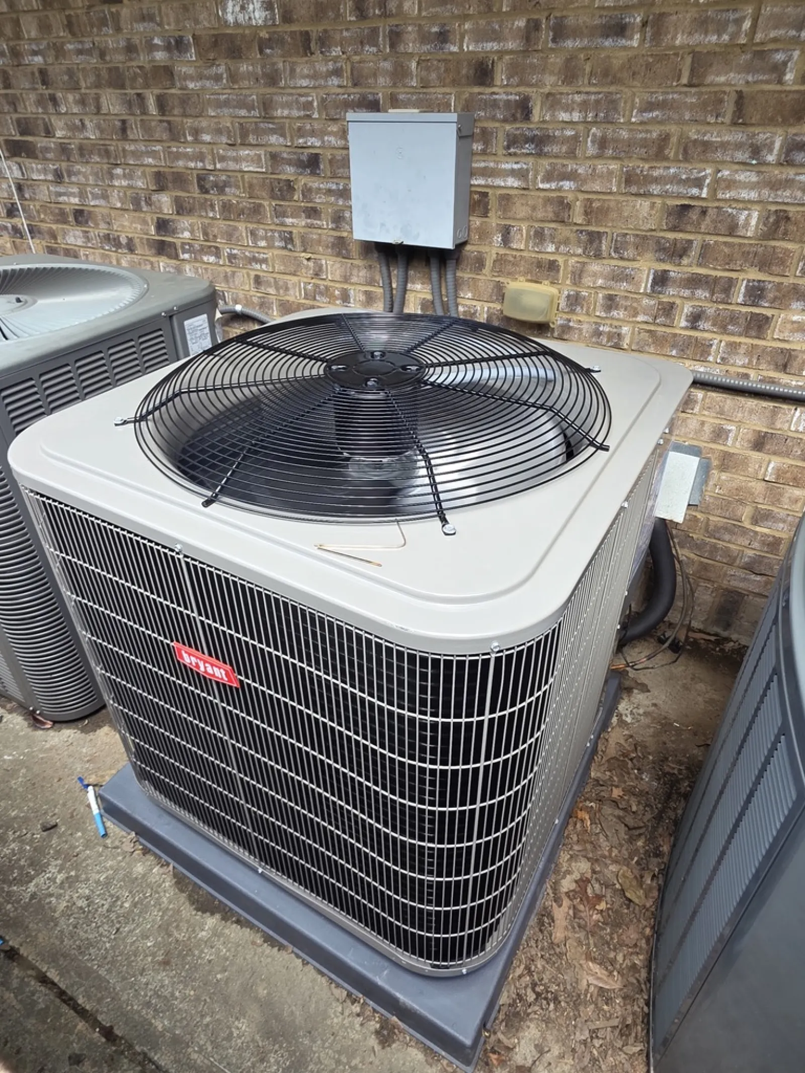 Outdoor HVAC air conditioning unit with a large top fan and metal casing next to a brick wall.