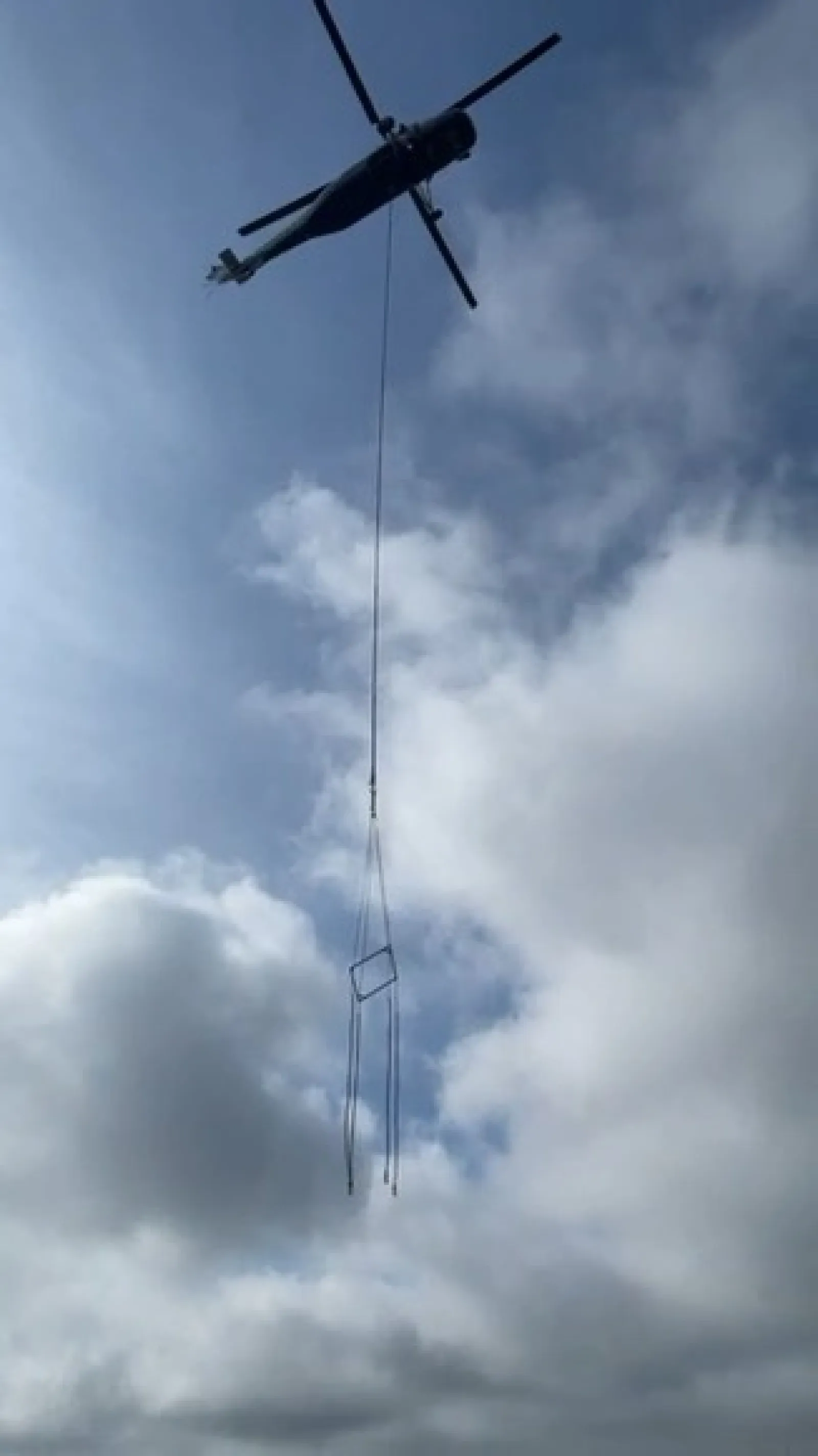 Helicopter lifts a large metal frame against a cloudy sky during daytime operation.