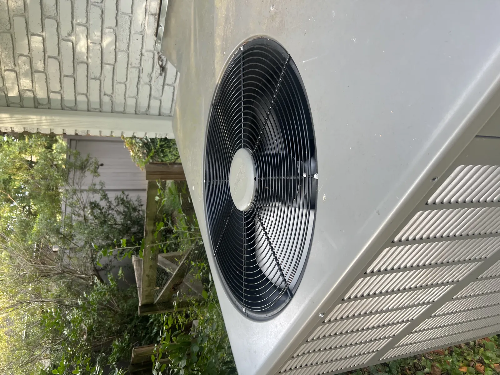 Outdoor air conditioning unit with large black fan and metal casing installed beside a brick wall.