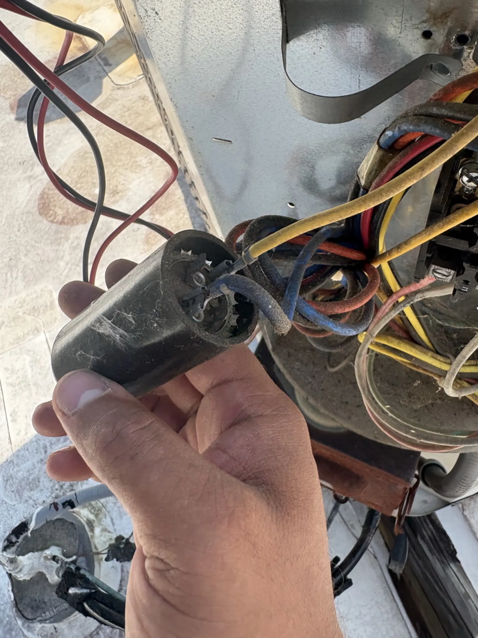 Hand holding a black electrical capacitor with exposed and connected multicolored wires in a mechanical unit.