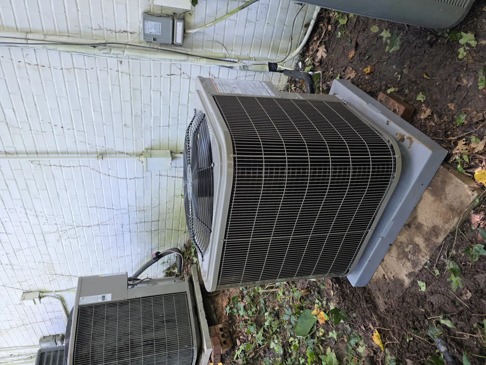 Two outdoor air conditioning units installed next to a white brick wall on a grassy patch with leaves.