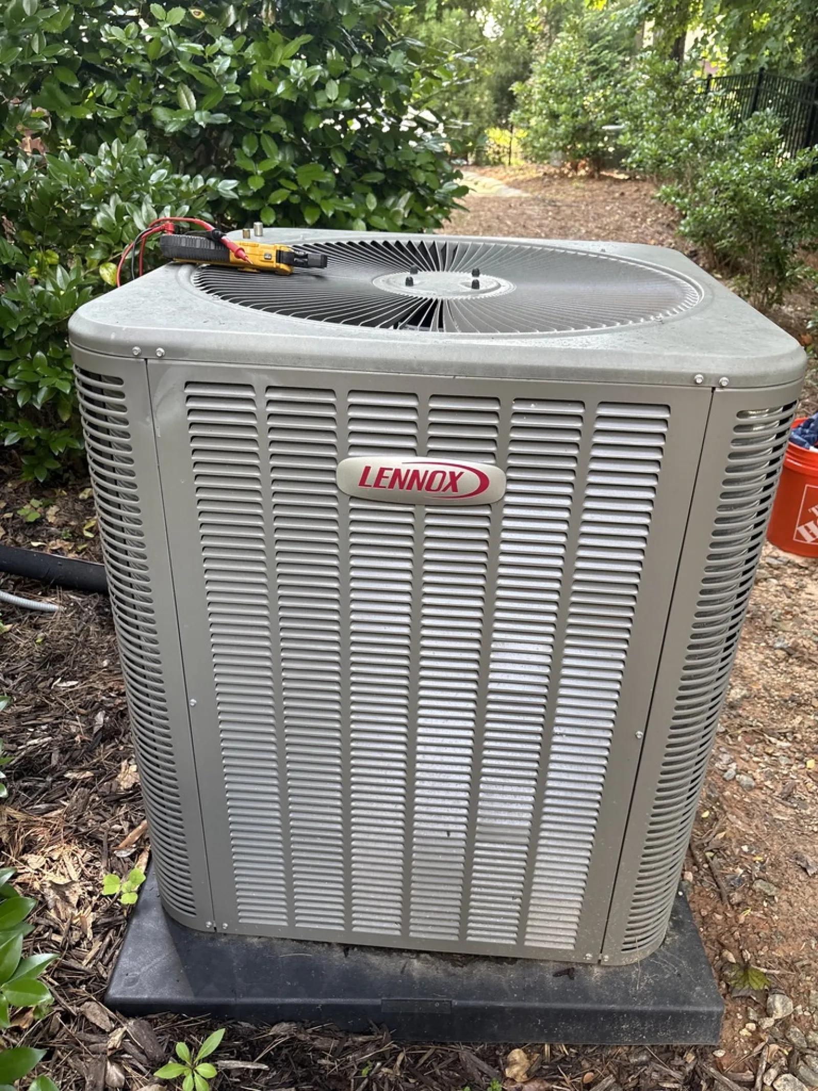 Outdoor Lennox air conditioning unit installed on a black plastic base surrounded by greenery and mulch.