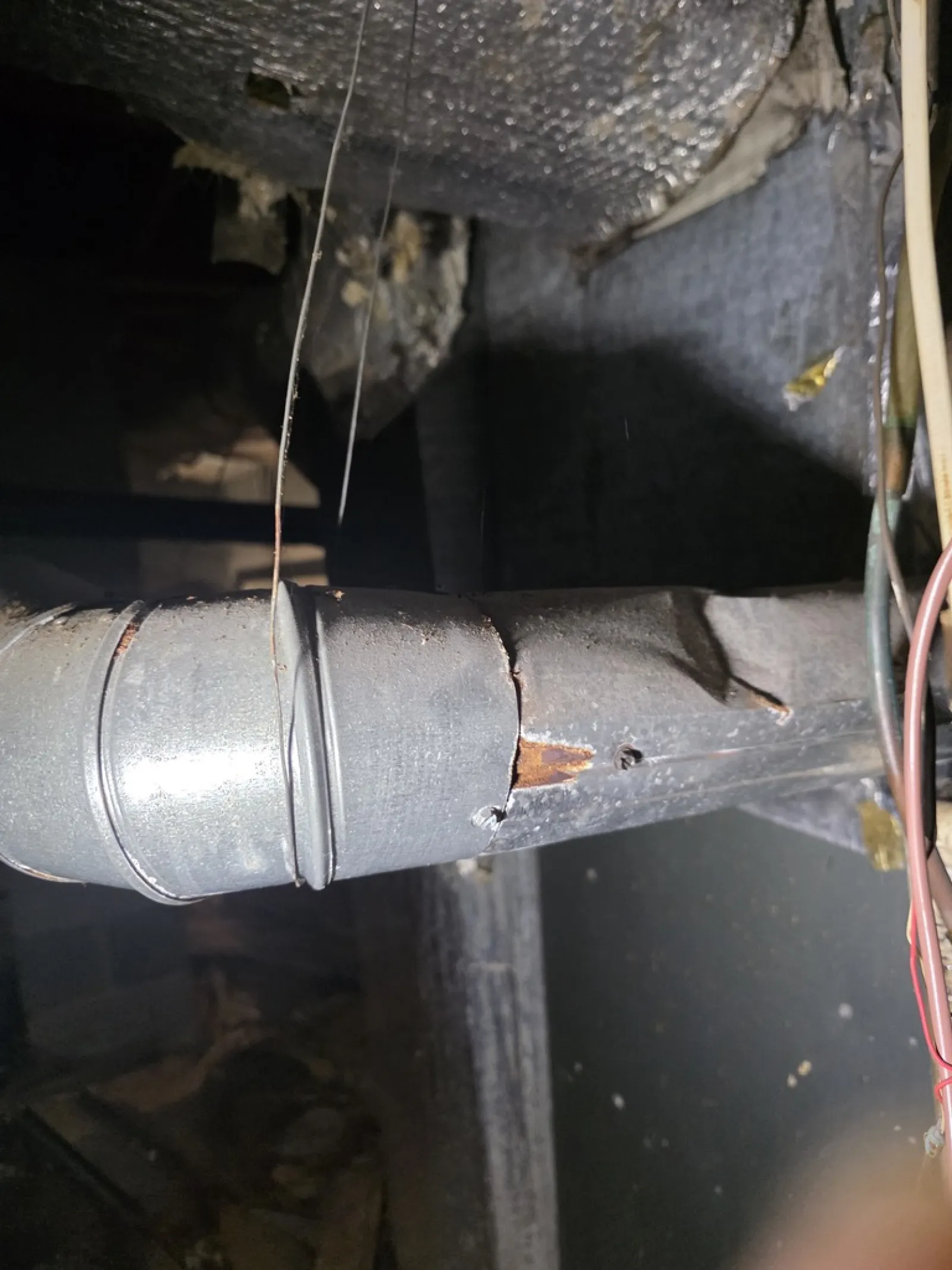 Rusty and cracked metal duct pipe in a dimly lit attic with insulation and wiring nearby.