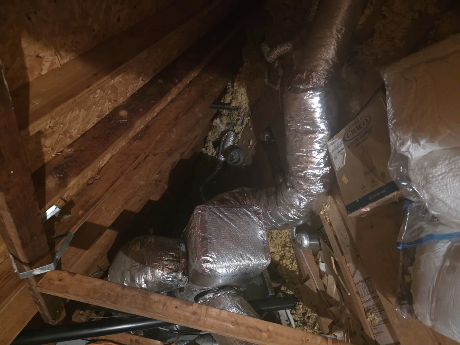 Insulated HVAC ductwork installed in a wooden attic with stored boxes and insulation materials.