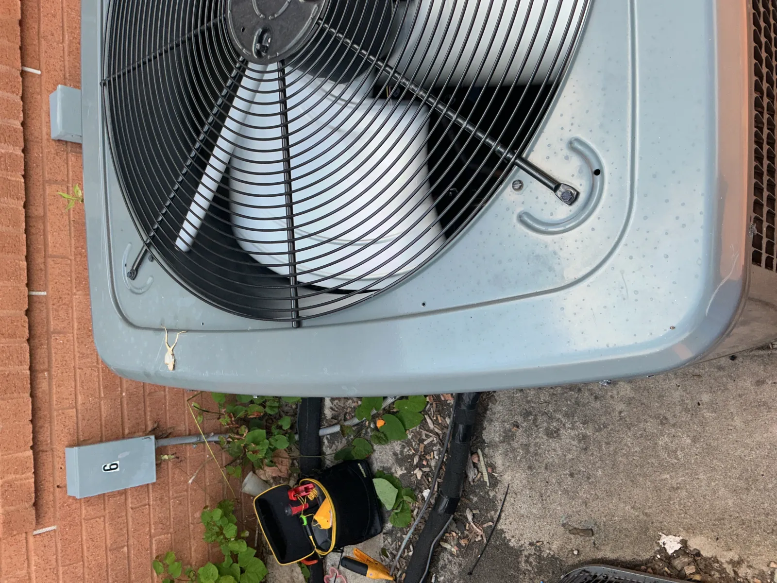 Outdoor air conditioning unit with protective fan grill near brick wall and electrical equipment on concrete