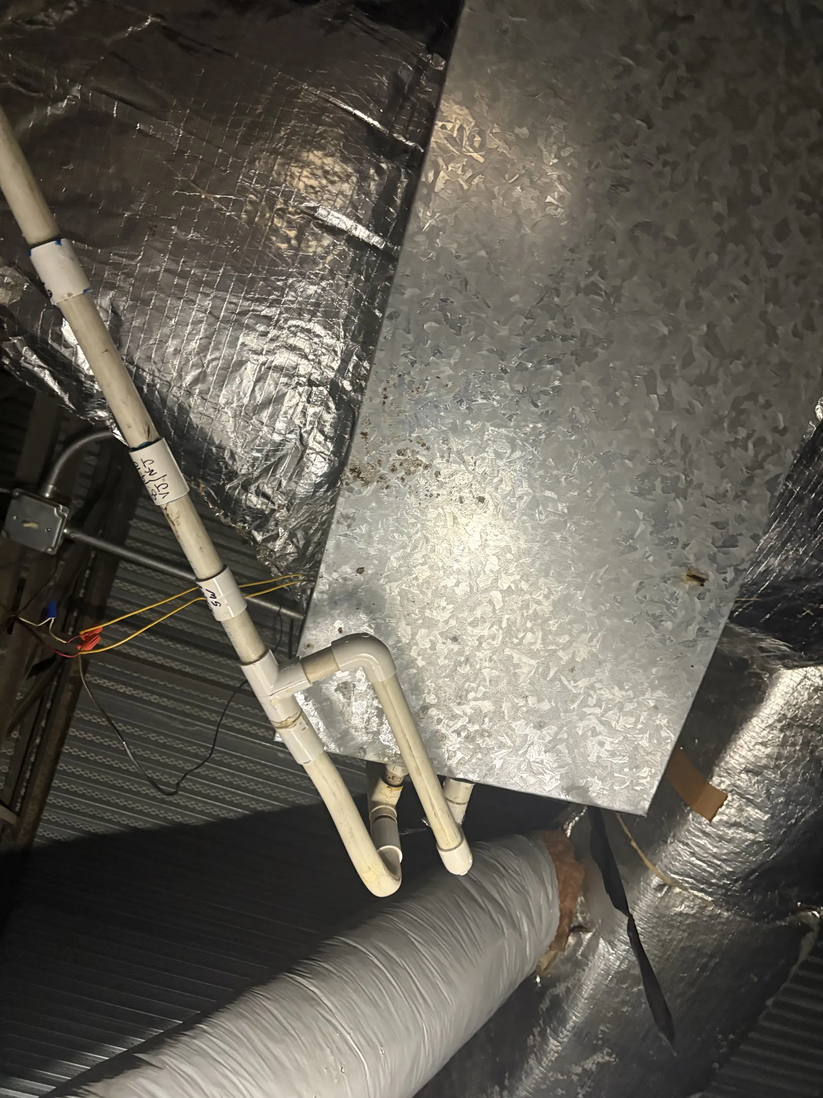 PVC pipes and insulated ductwork connected to a galvanized metal HVAC unit in an industrial ceiling space.