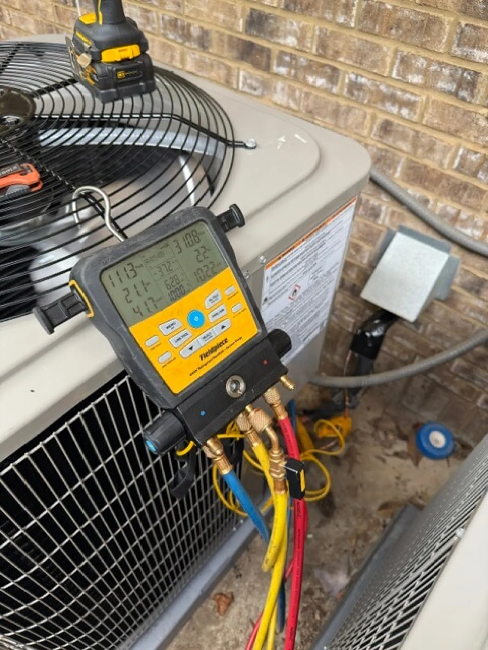 Digital manifold gauge connected to an outdoor HVAC unit with colored hoses and tools nearby.