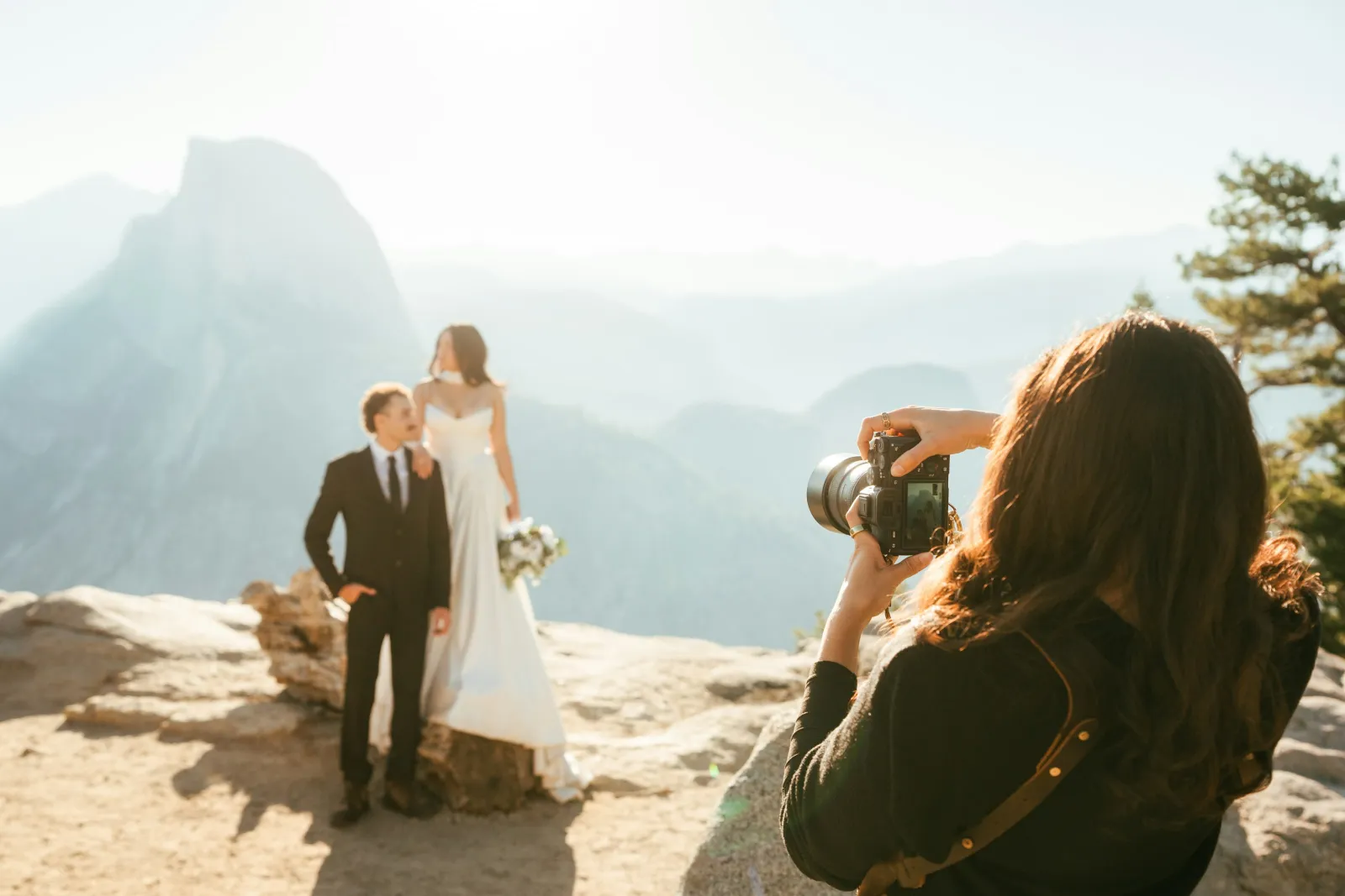 Photographer capturing a bride and groom on a mountain cliff with scenic valley and peaks in the background.