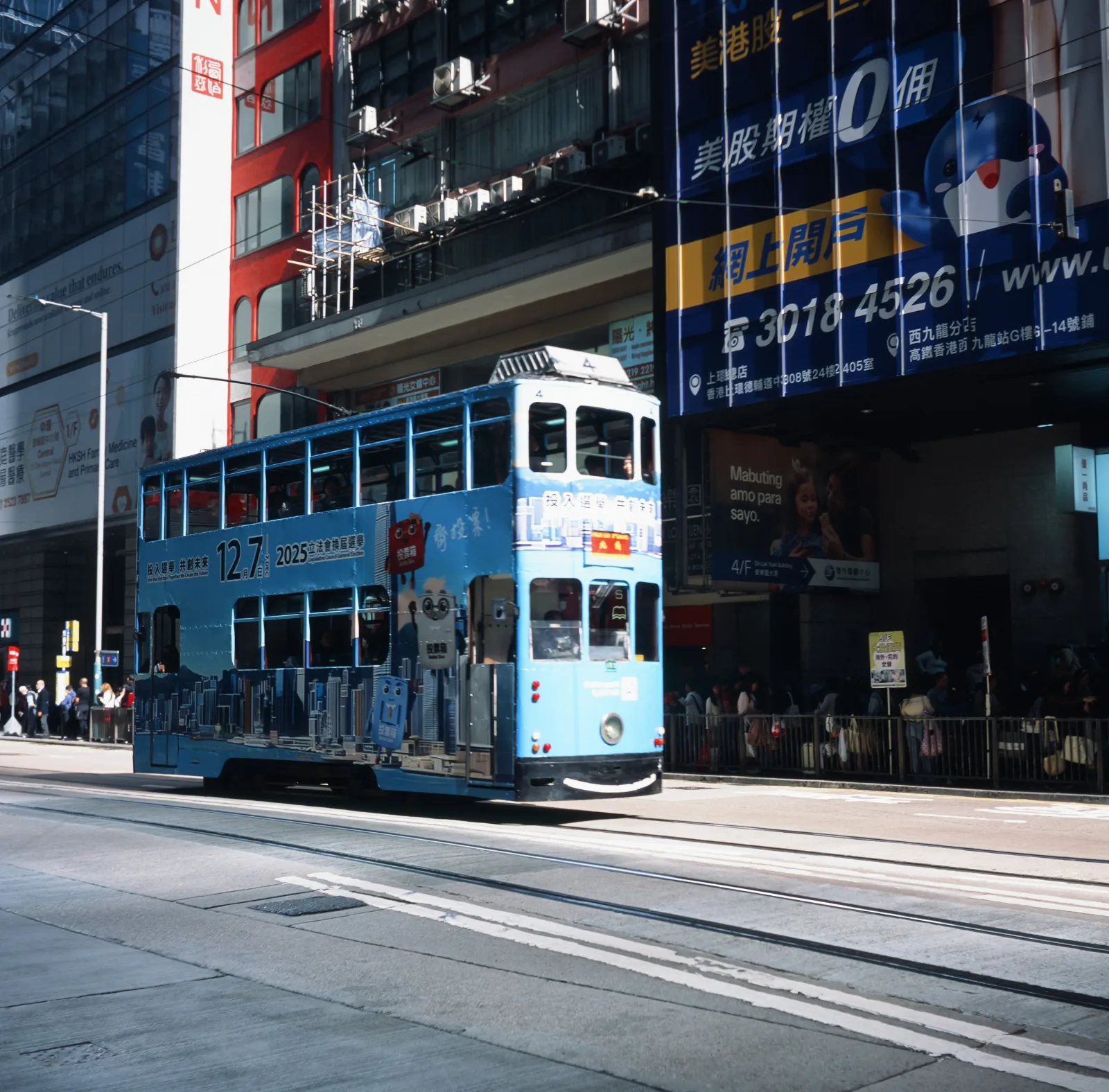 Blue double-decker tram on city street with advertisements and tall buildings in background.