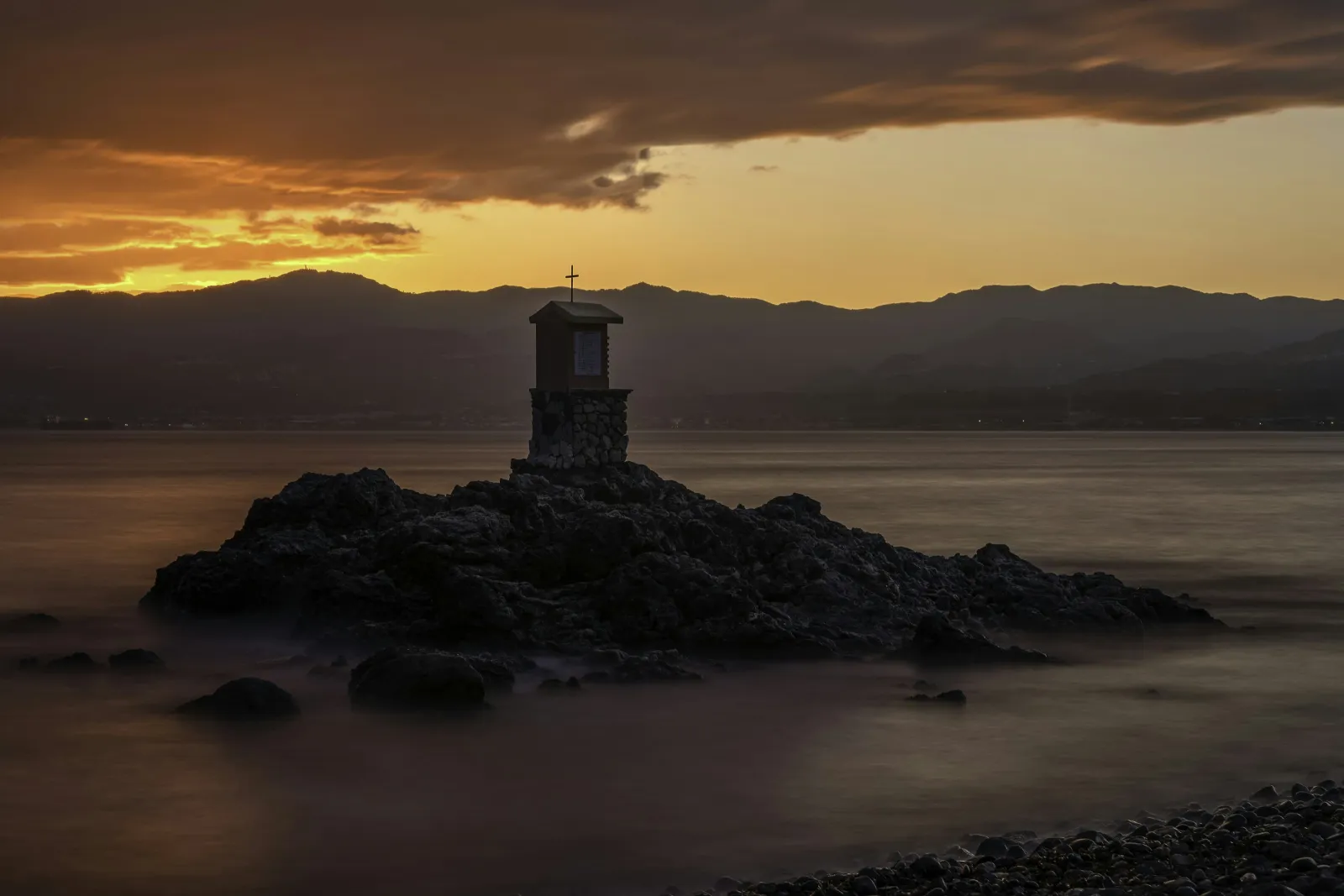 Small stone chapel with cross on rocky island at sunset with mountains and cloudy sky in the background