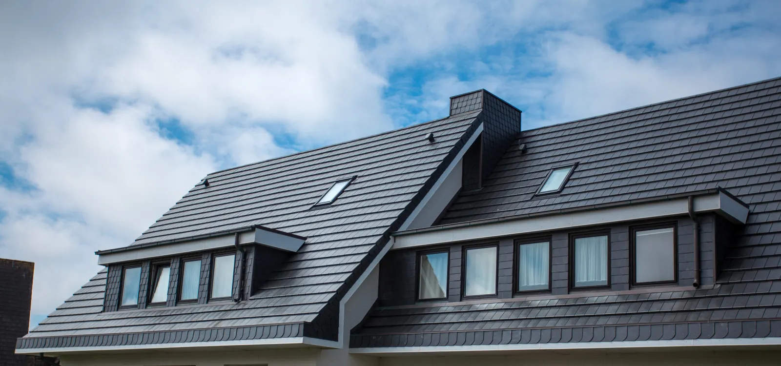 Modern house roof with stylish shingles under a blue cloudy sky, showcasing architectural design.