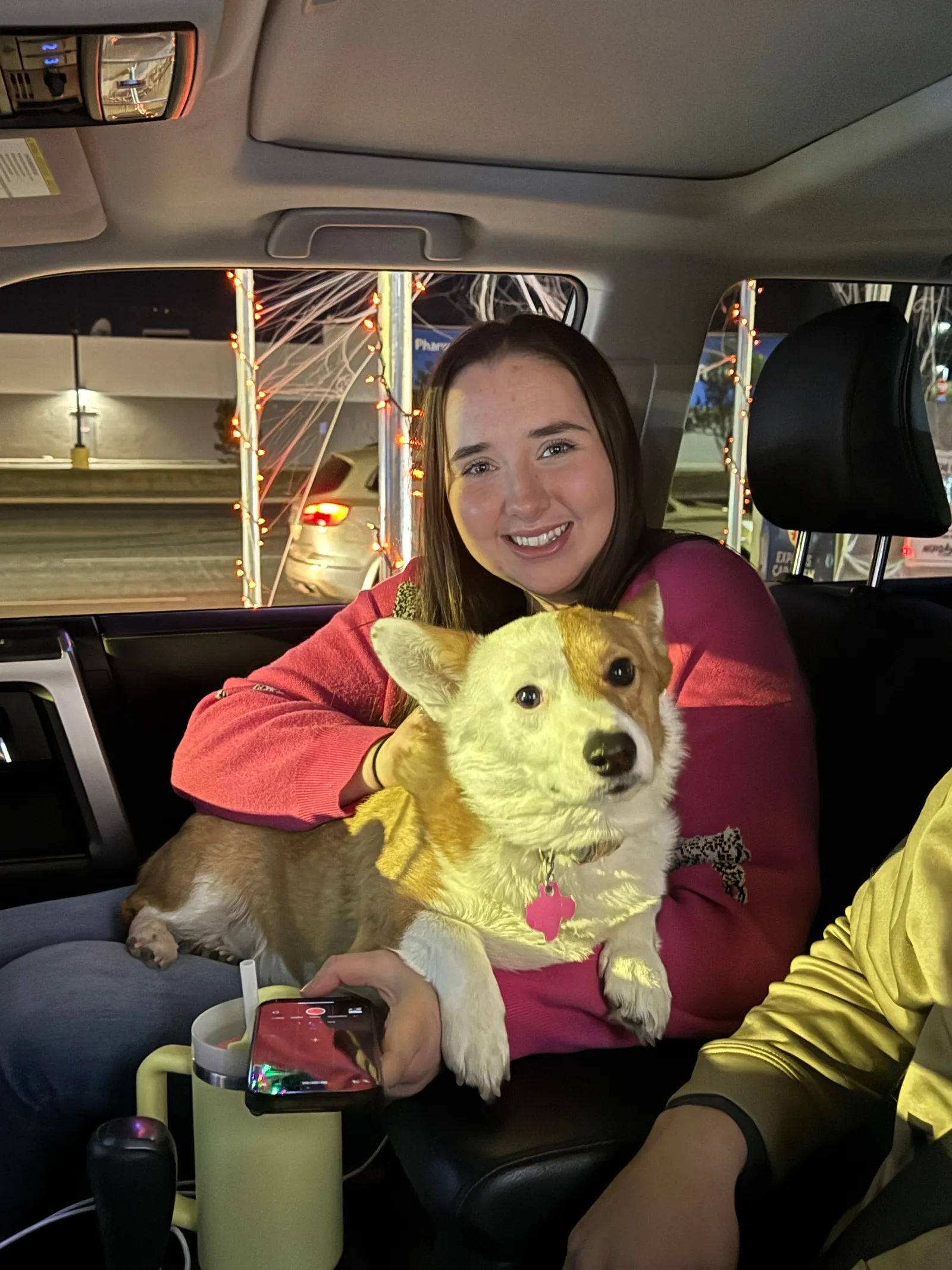 Smiling woman in car holding a corgi dog with a pink tag, phone, and night street lights in background