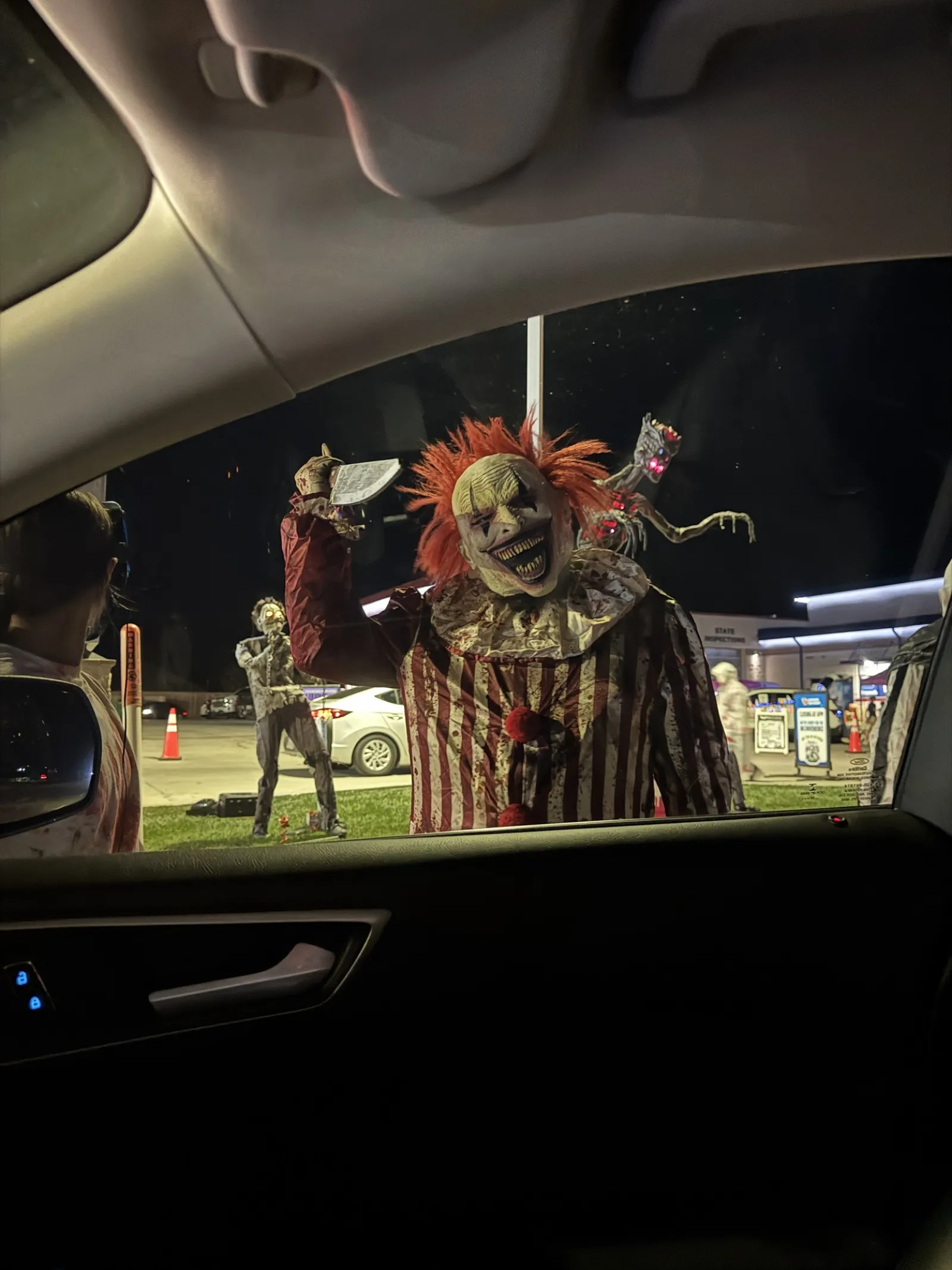Scary clown with red hair and striped costume holding a cleaver outside a car at night during Halloween event.