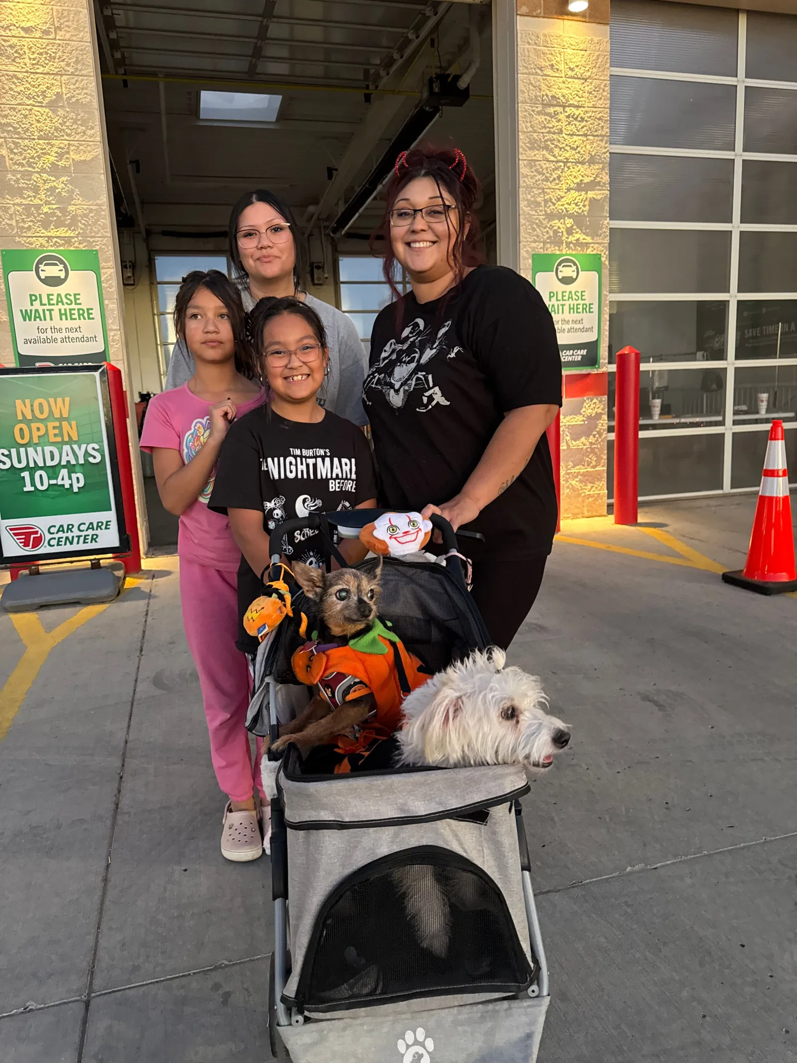 Group of four smiling people with two dogs in a stroller outside a car care center at sunset.