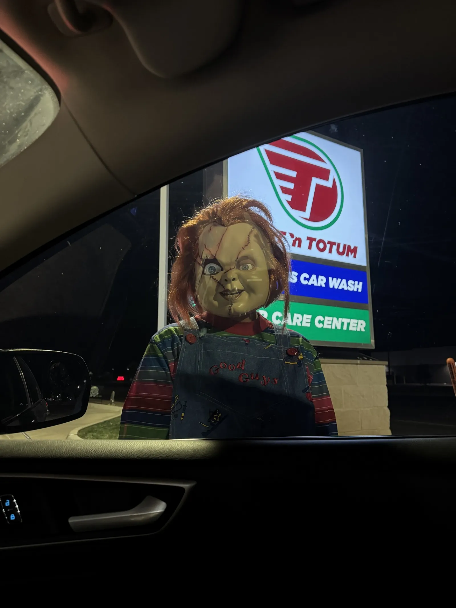 Chucky doll with red hair and overalls standing outside a car window at night near a gas station sign.